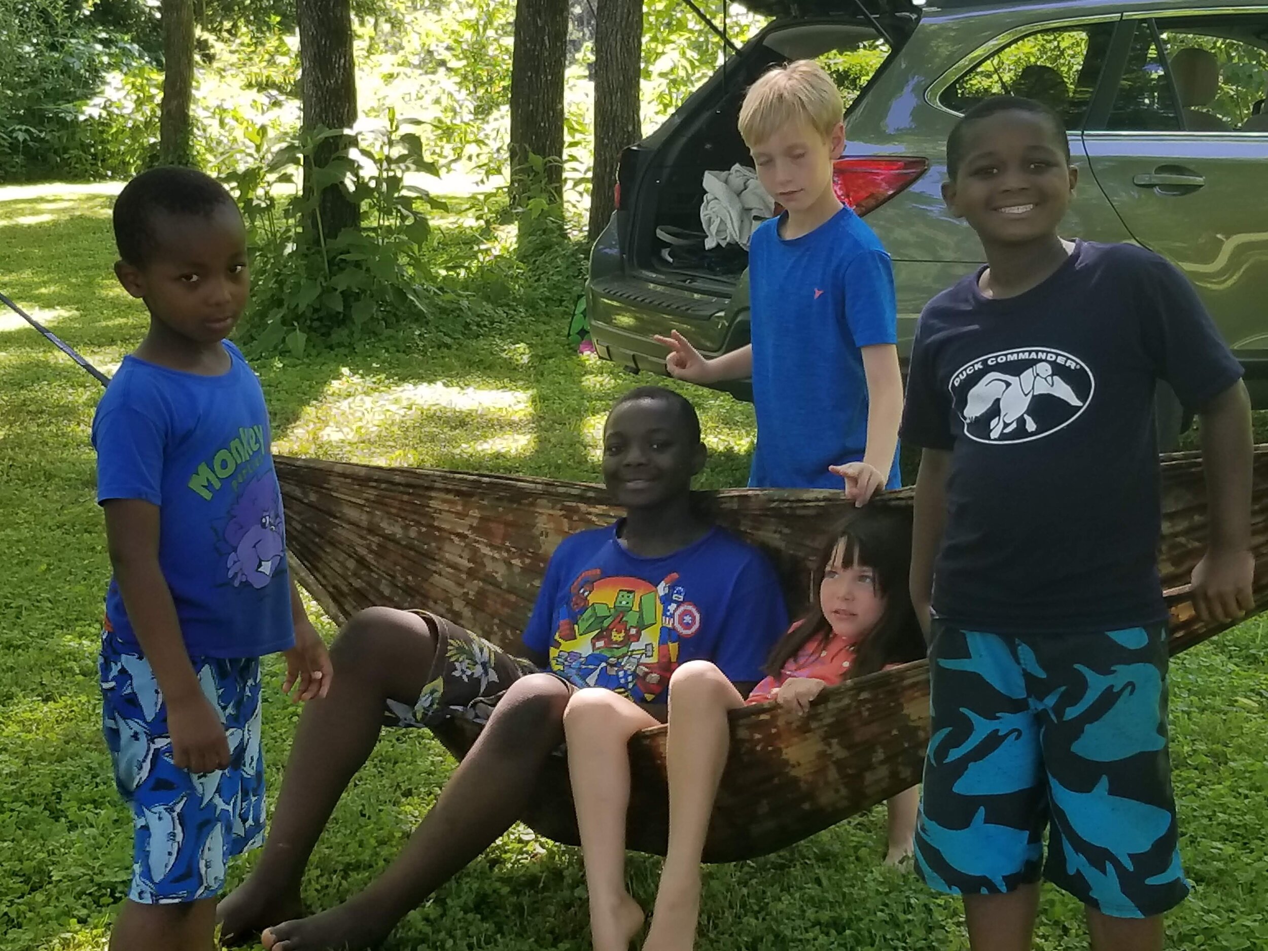 Group of children outdoors near a hammock and parked car, with trees and grass in the background.