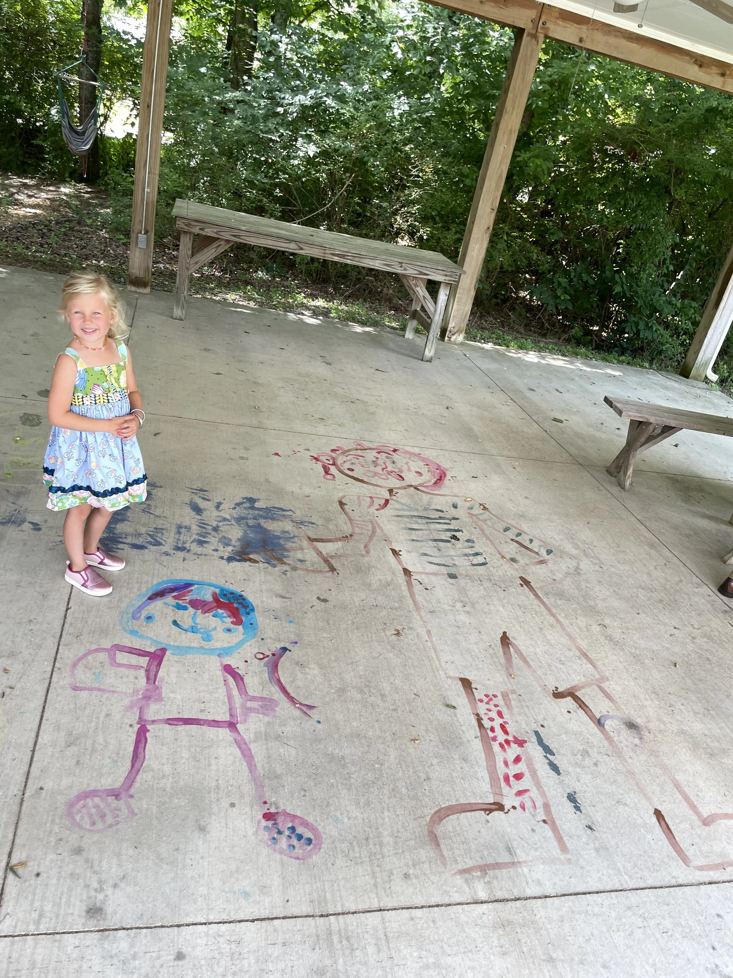 A child in a colorful dress and pink sandals stands on a concrete surface, smiling. Next to her on the ground are large, colorful chalk drawings of two people. The background features green foliage and two wooden benches under a pavilion.