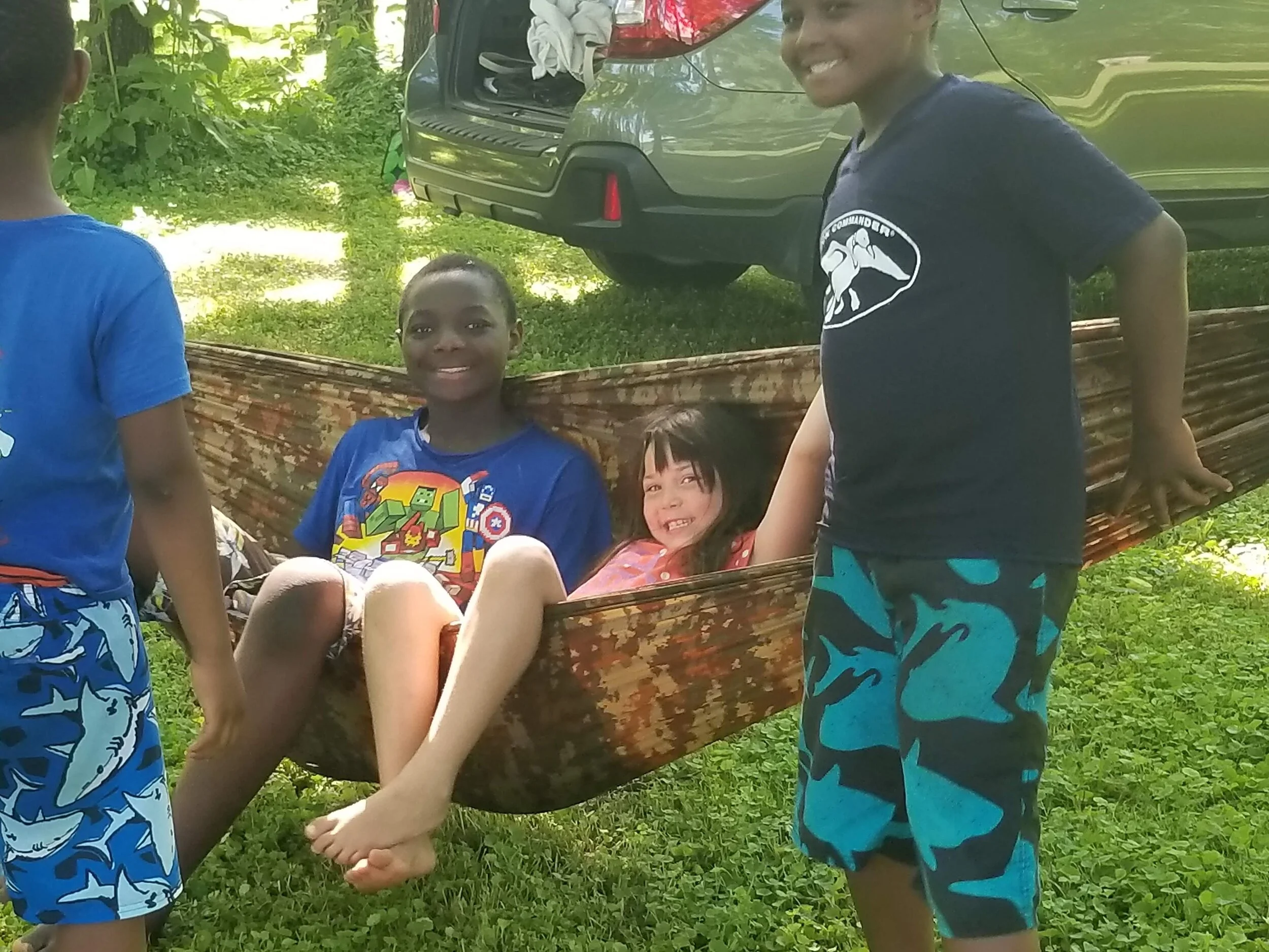 Children playing in a hammock outdoors next to a car.