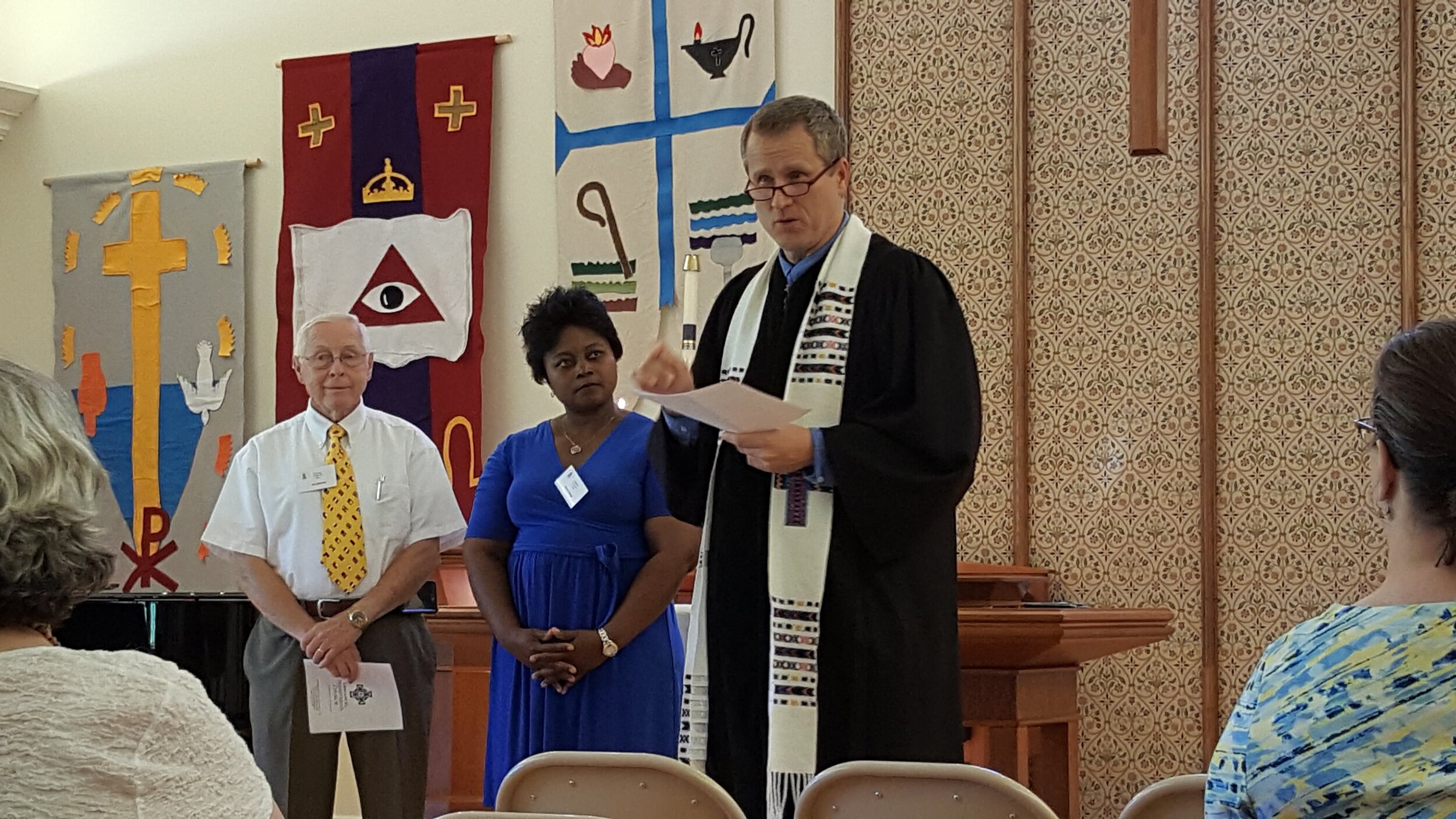 Three people stand in a church setting, with religious banners in the background. A man wearing a clerical robe and stole speaks, holding papers. A woman in a blue dress and a man in a white shirt and yellow tie stand beside him. An audience is visib