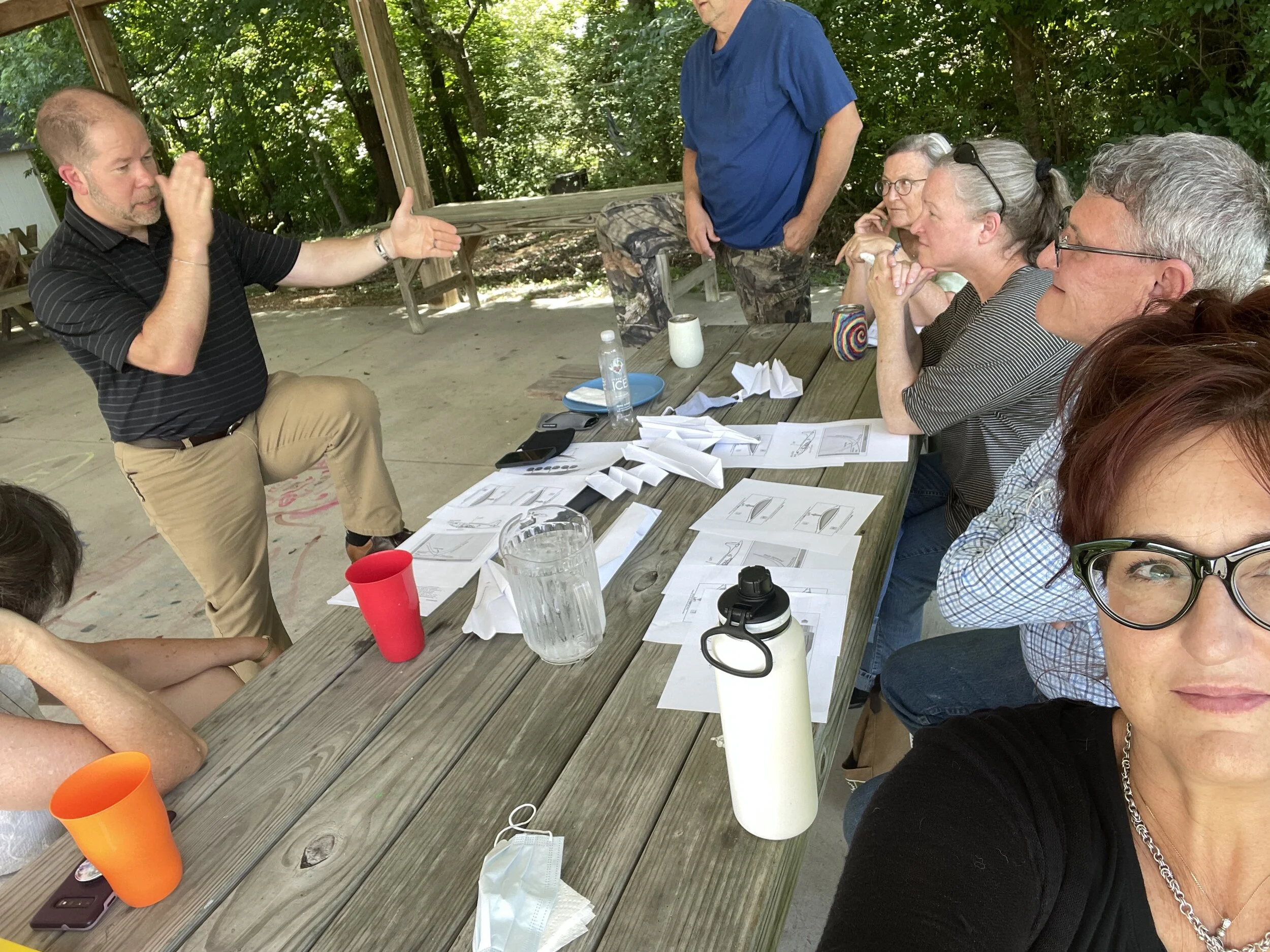 A group of people in a casual outdoor meeting, sitting on picnic tables with papers and cups scattered on the table. One man stands and gestures while others are seated, listening attentively. They are in a shaded, wooded area with some wearing casua