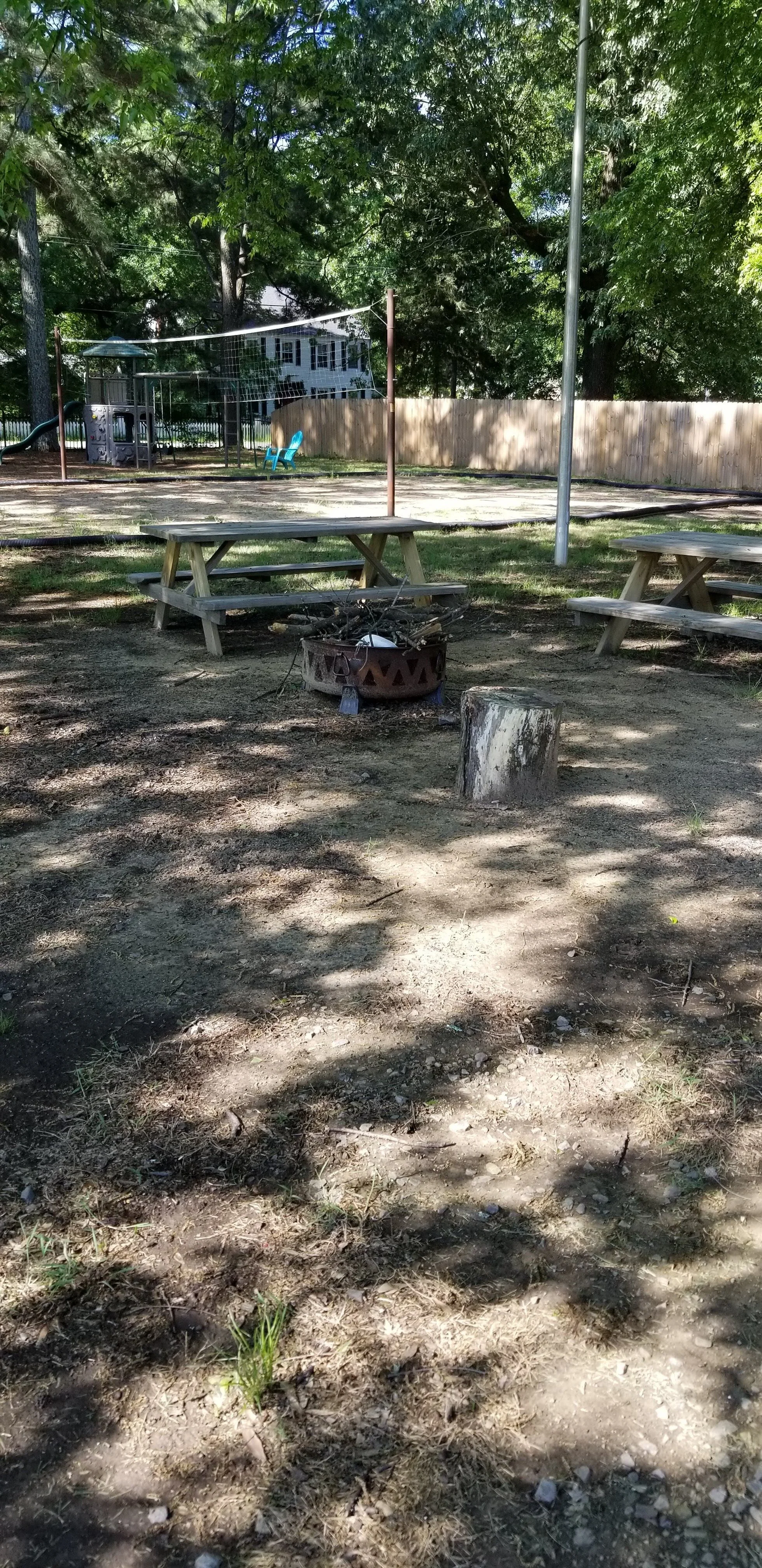 Outdoor scene with picnic tables and a fire pit, surrounded by trees, a wooden fence, and a residential house in the background. A playground with a jungle gym and a blue chair are visible.
