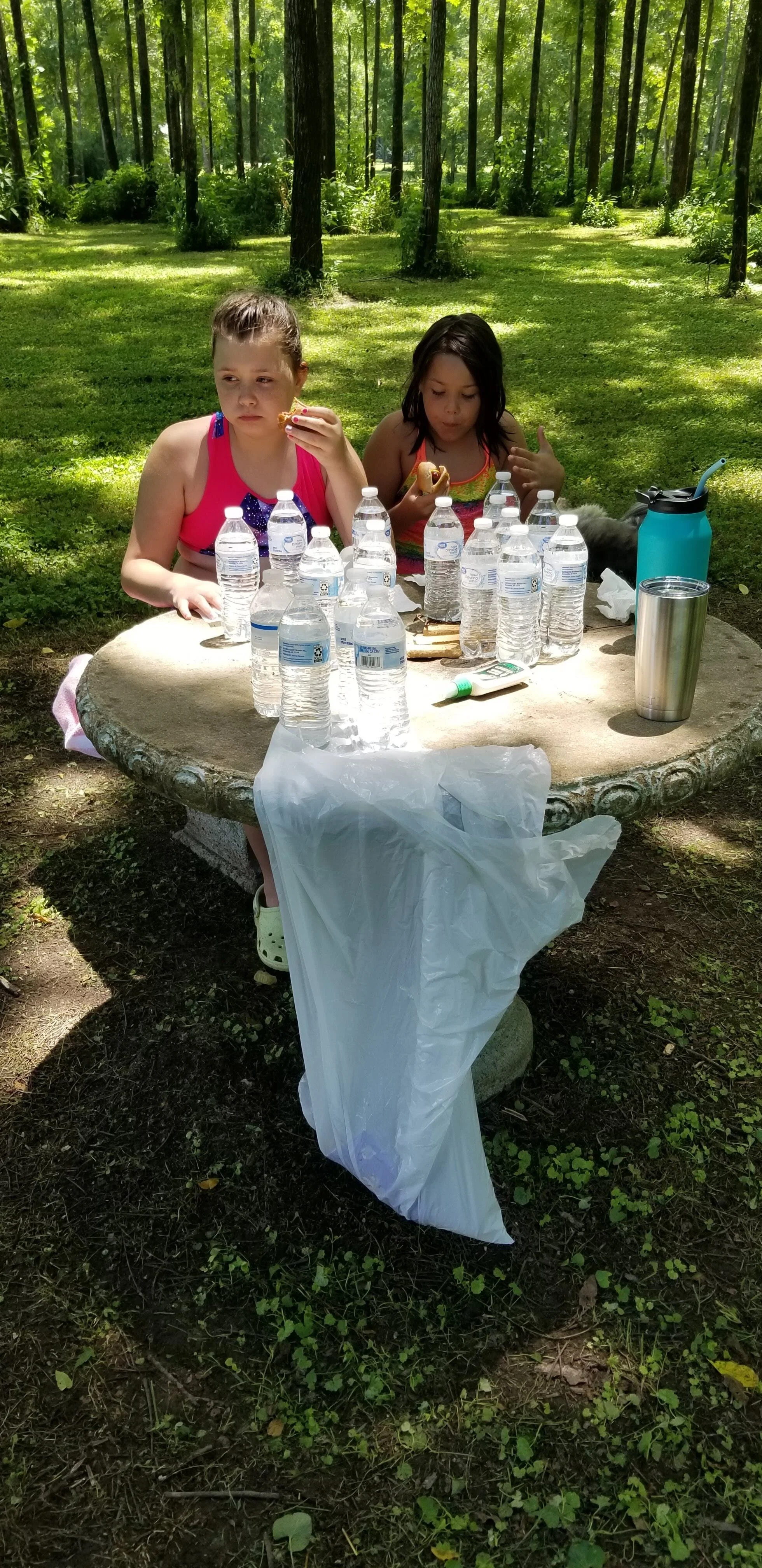 Two people sitting at a stone picnic table with bottled water, a plastic bag, and a thermos in a wooded area.