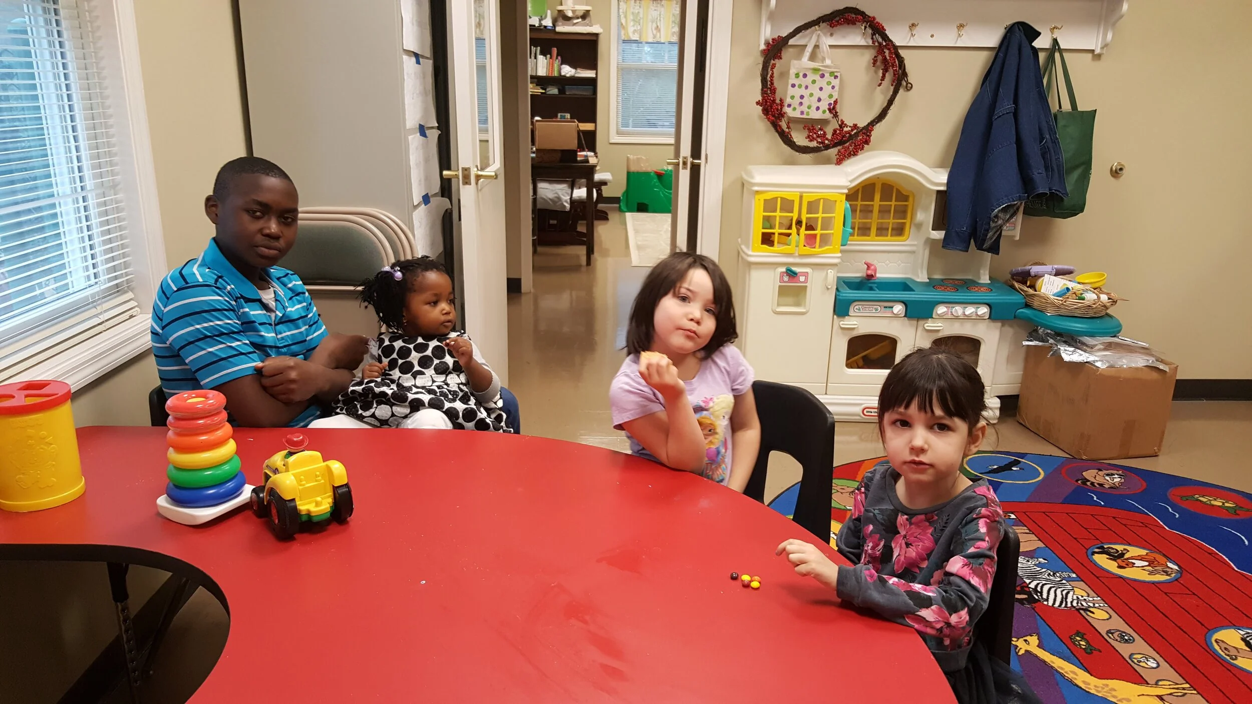 Four children sitting around a red table in a playroom with toys and play kitchen in the background.