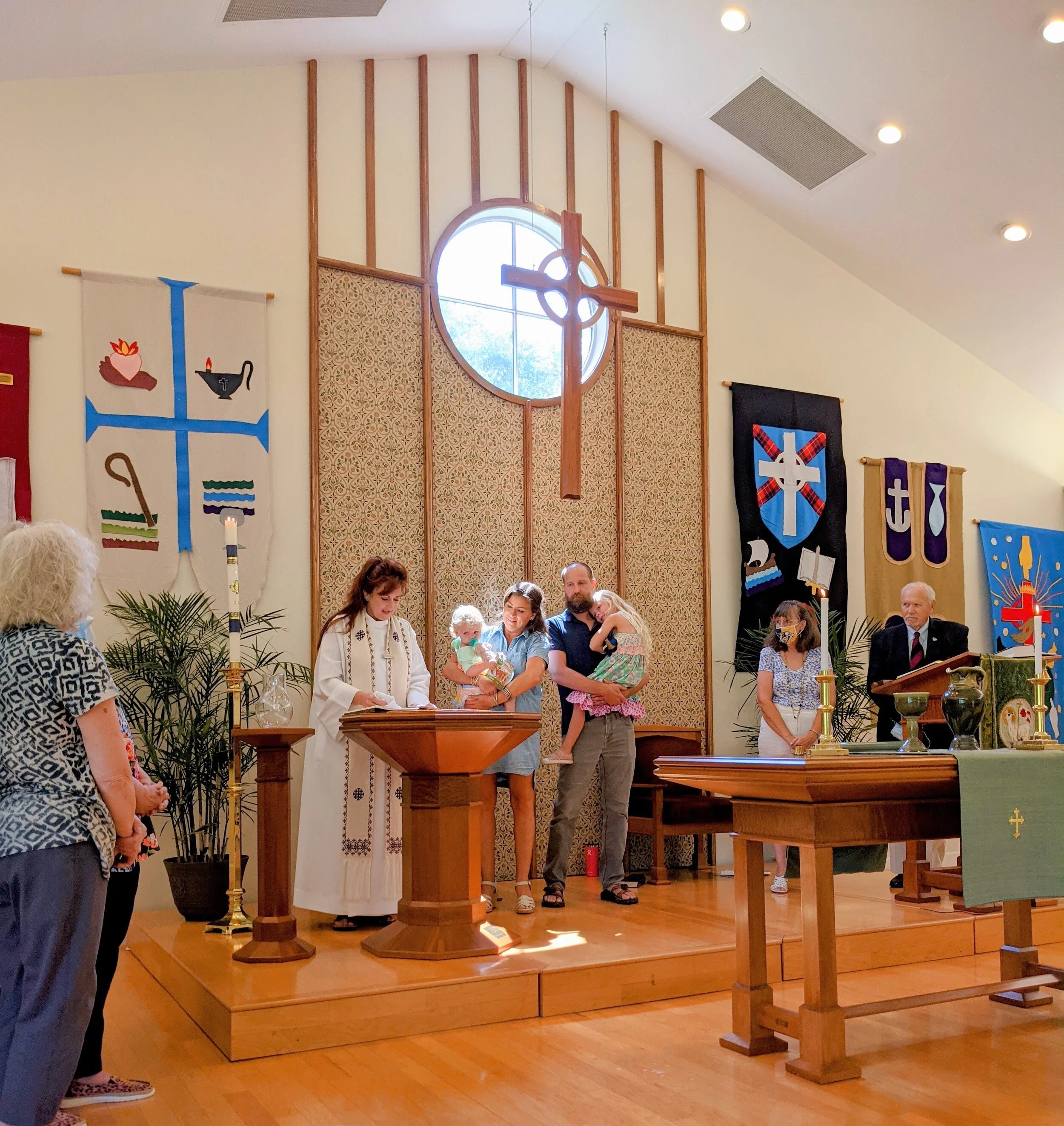 Church ceremony with people at altar, priest, banners, cross sculpture, family holding children, and candles.