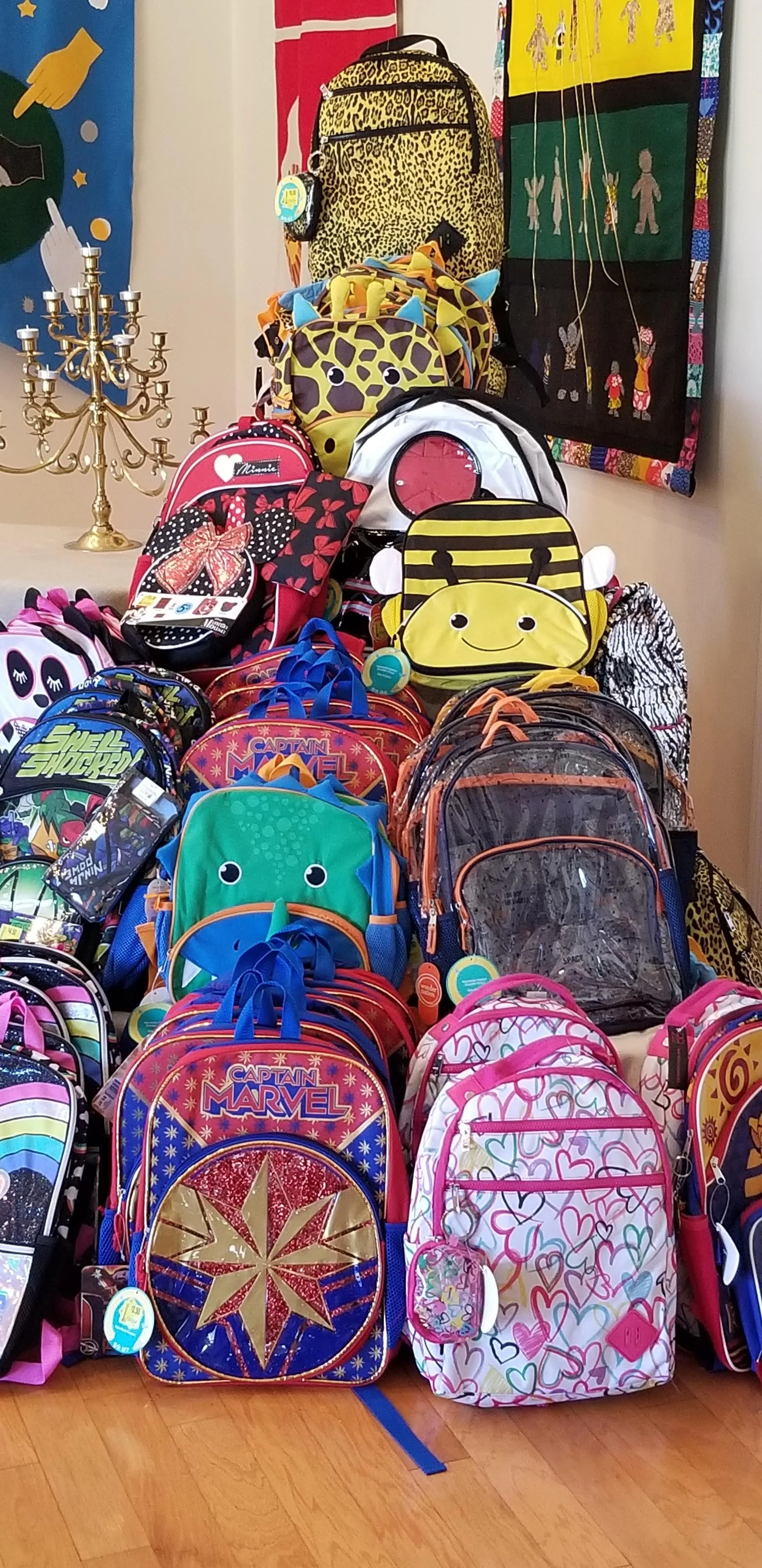 A variety of colorful children's backpacks stacked on a wooden floor. Designs include animal faces, hearts, and popular characters. A candelabrum and decorative wall hangings are visible in the background.
