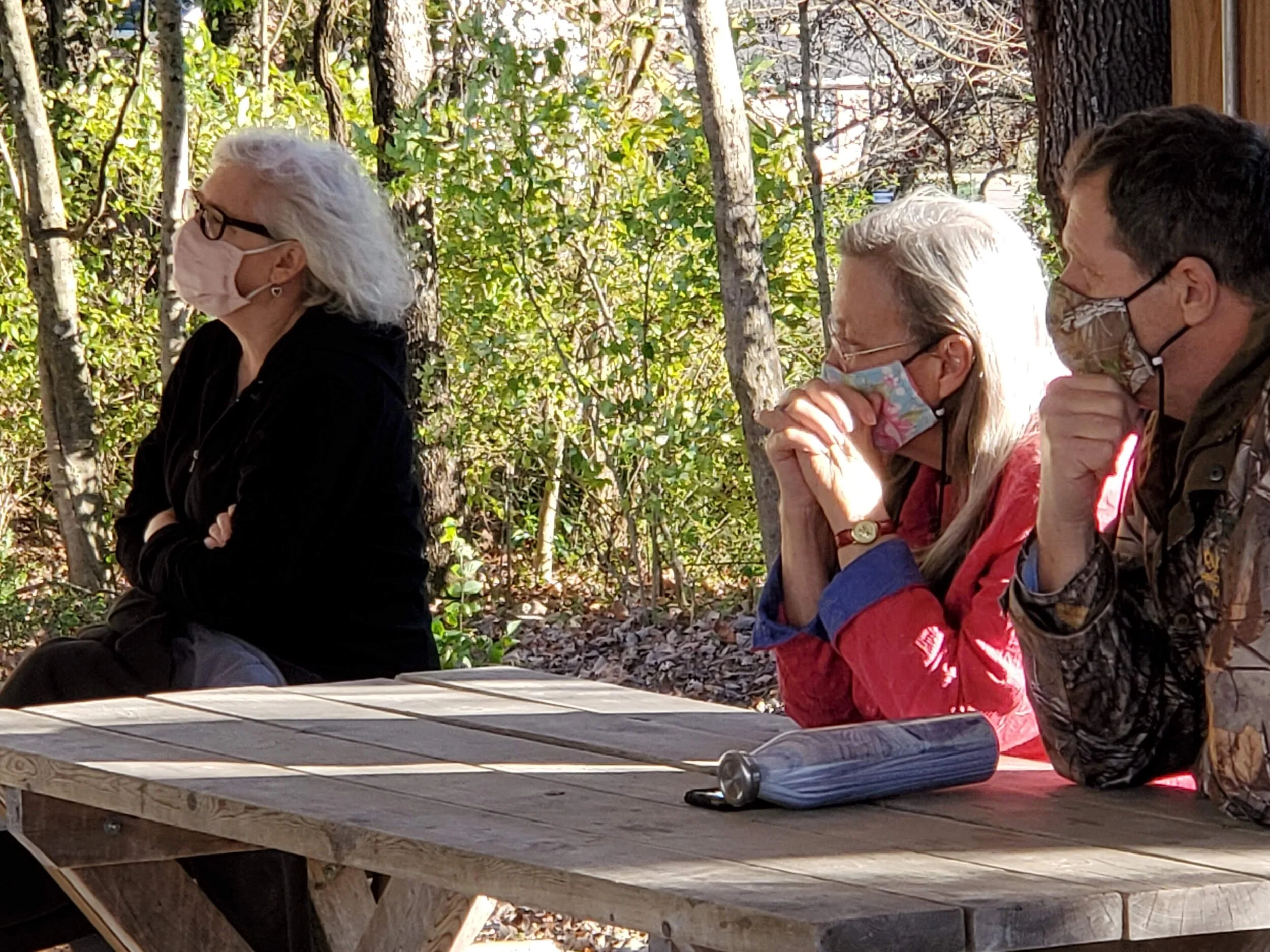 Three people wearing face masks sitting at a wooden picnic table outdoors, surrounded by trees.