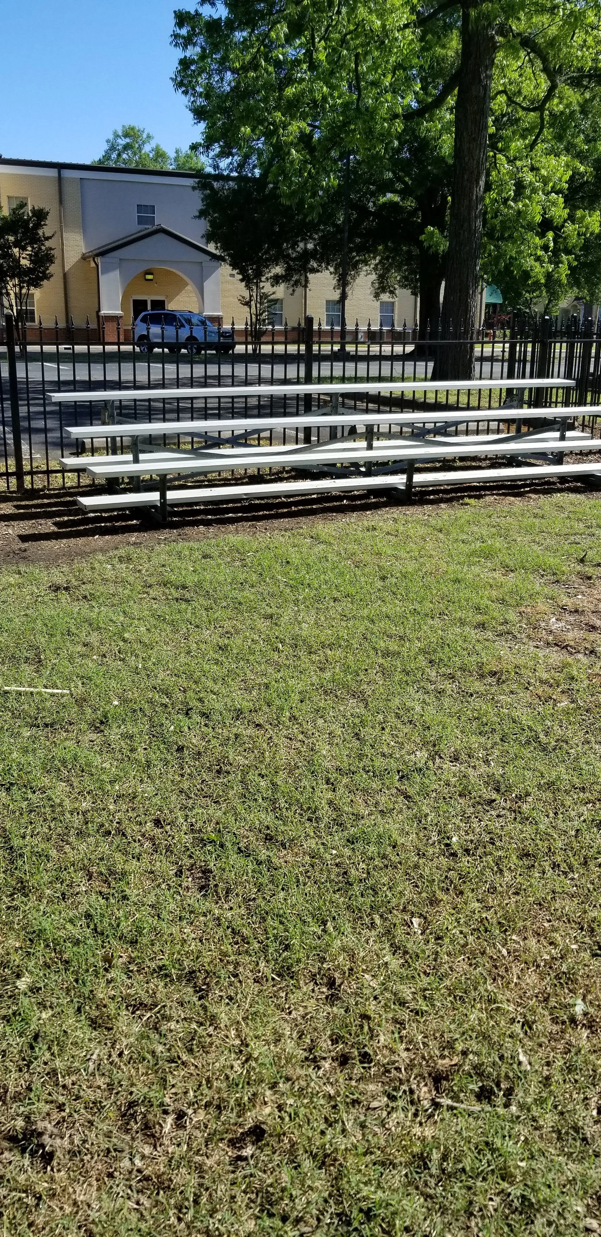 Bleachers in a grassy area with a fence and building in the background, trees providing shade, and a parked vehicle visible.