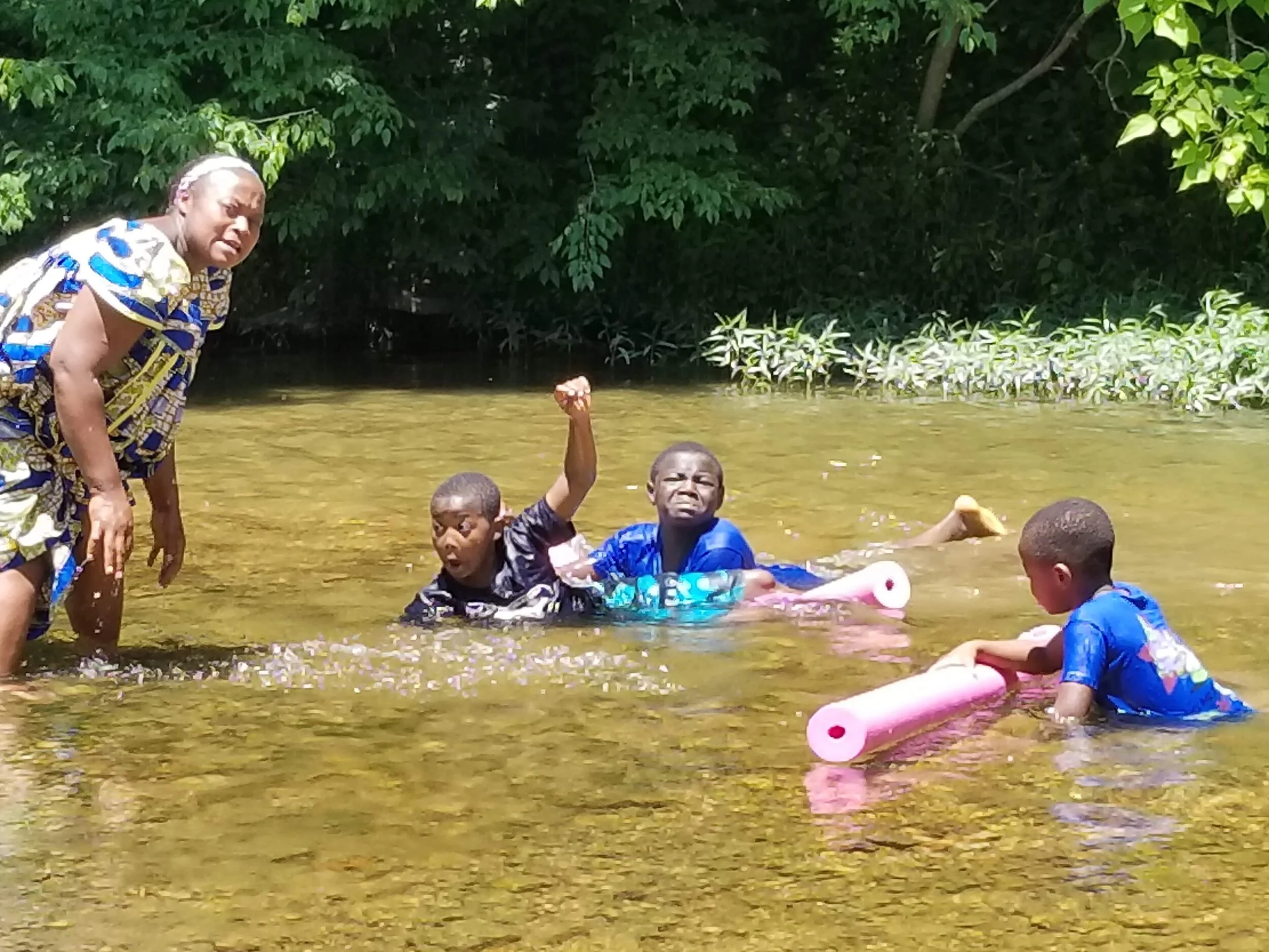 Group of children playing in a shallow creek with pool noodles and an adult nearby, surrounded by greenery.