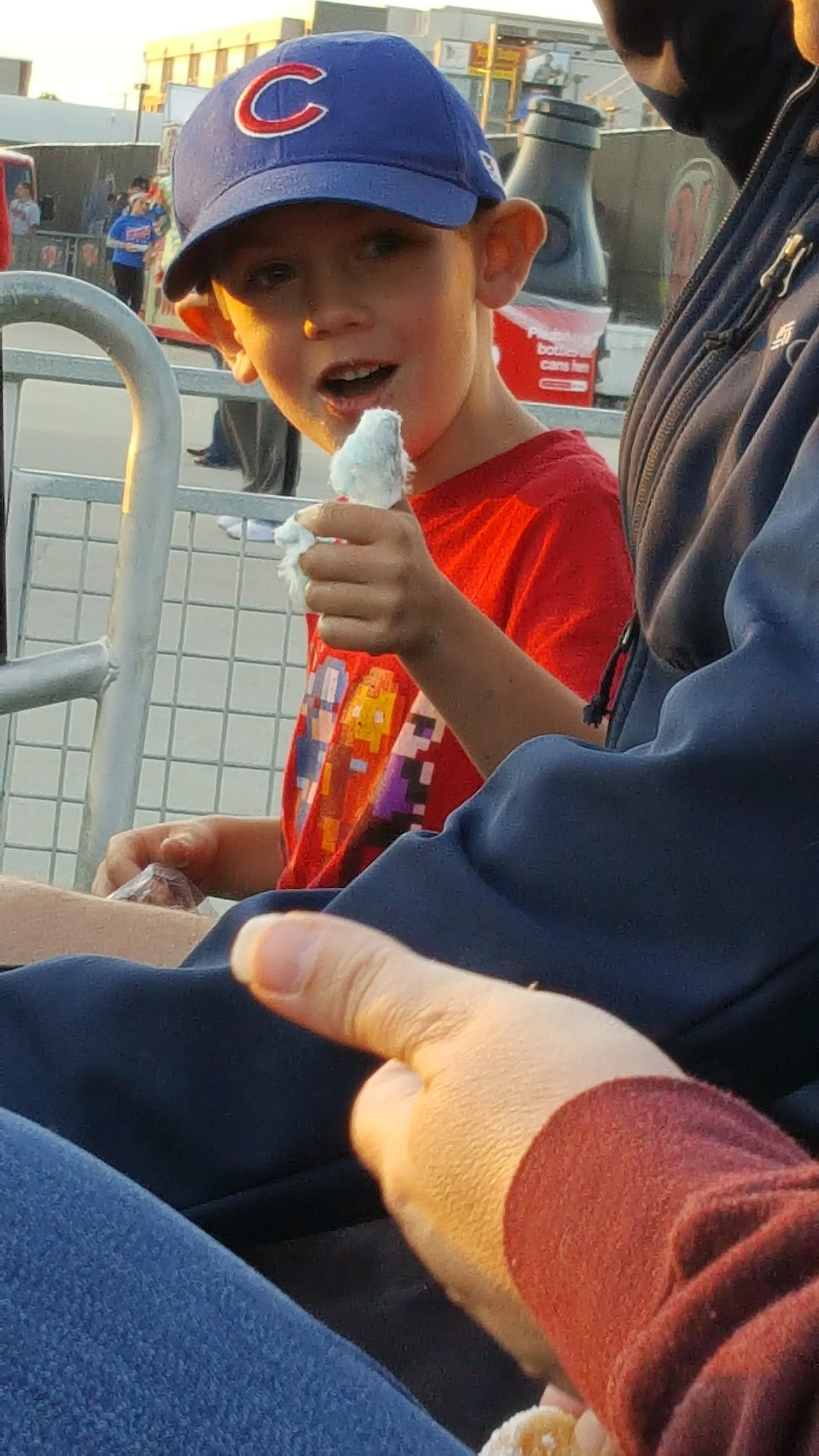 Child wearing a baseball cap and red shirt eating cotton candy at an outdoor event.