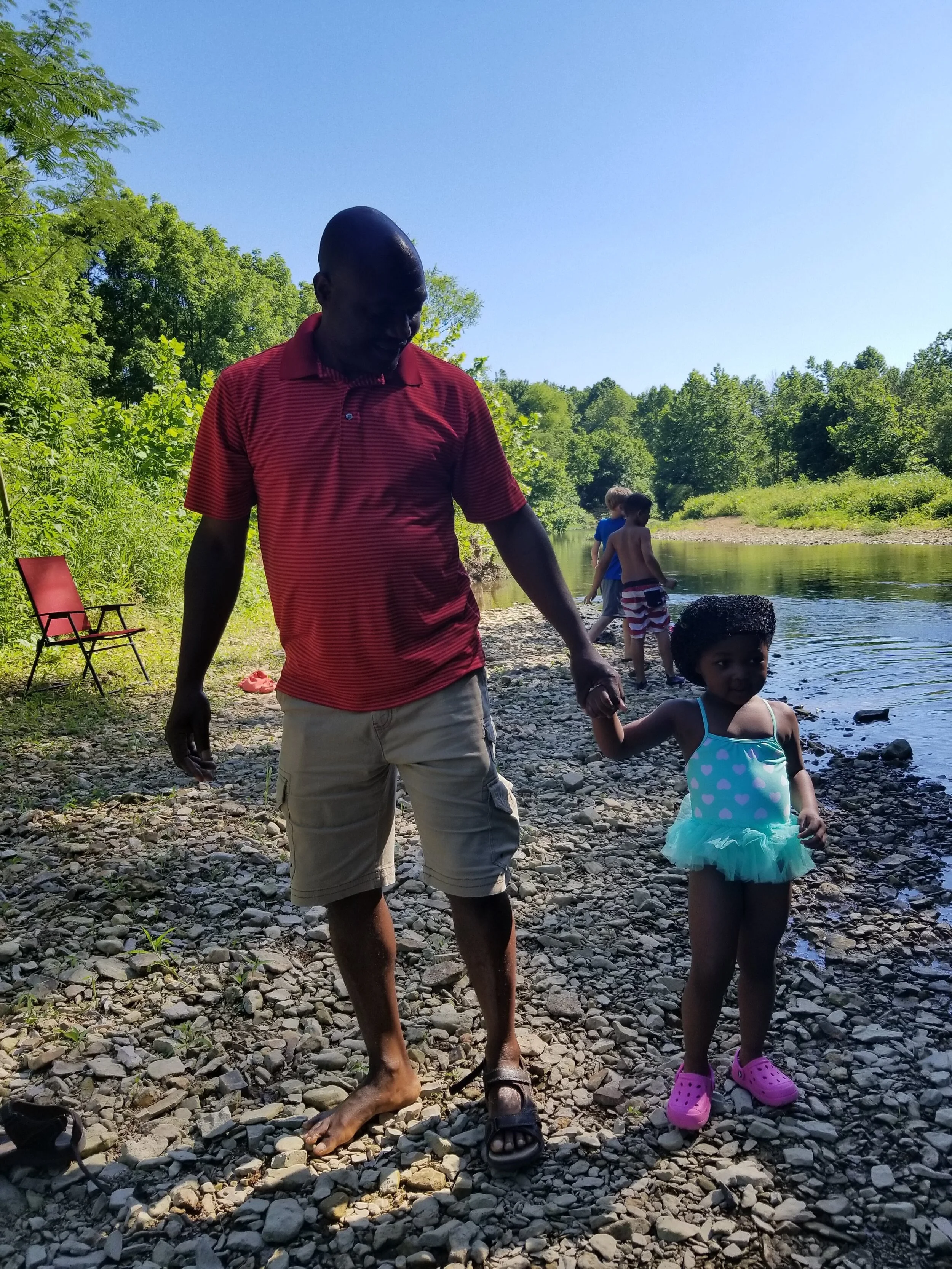 A man in a red shirt and beige shorts holding the hand of a young girl in a bathing suit standing by a rocky riverbank. Other children playing nearby. Bright, sunny day with green trees in the background.