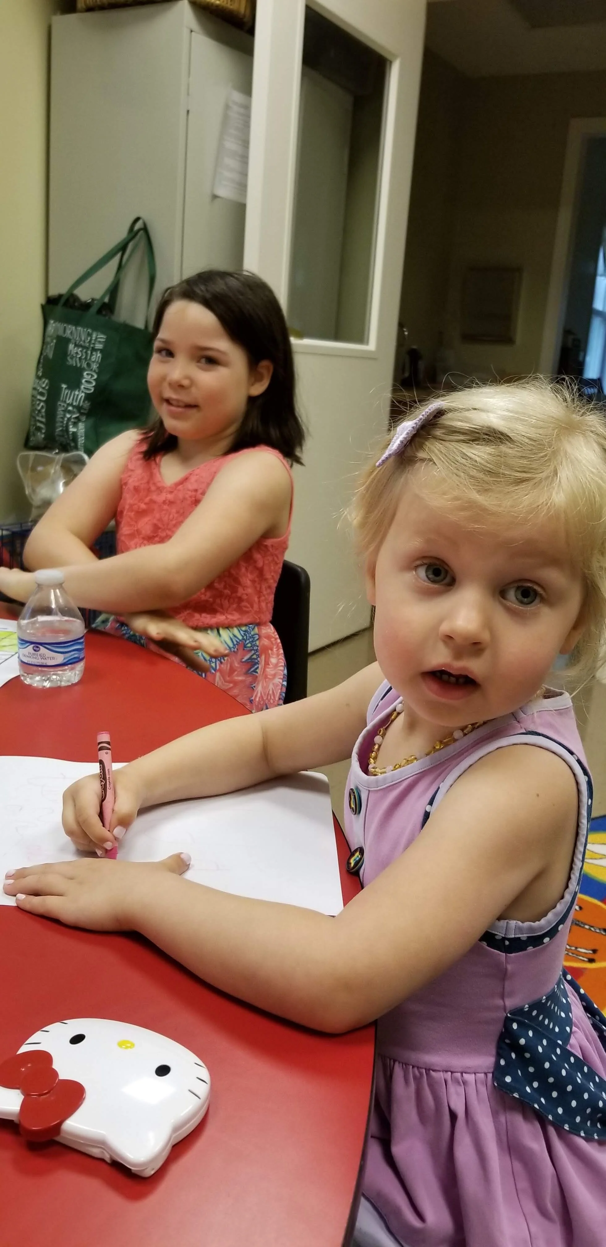 Two young girls sitting at a red table engaged in drawing. One is wearing a purple dress and holding a pink crayon, while the other has dark hair and wears a pink top. A water bottle and a Hello Kitty purse are on the table.