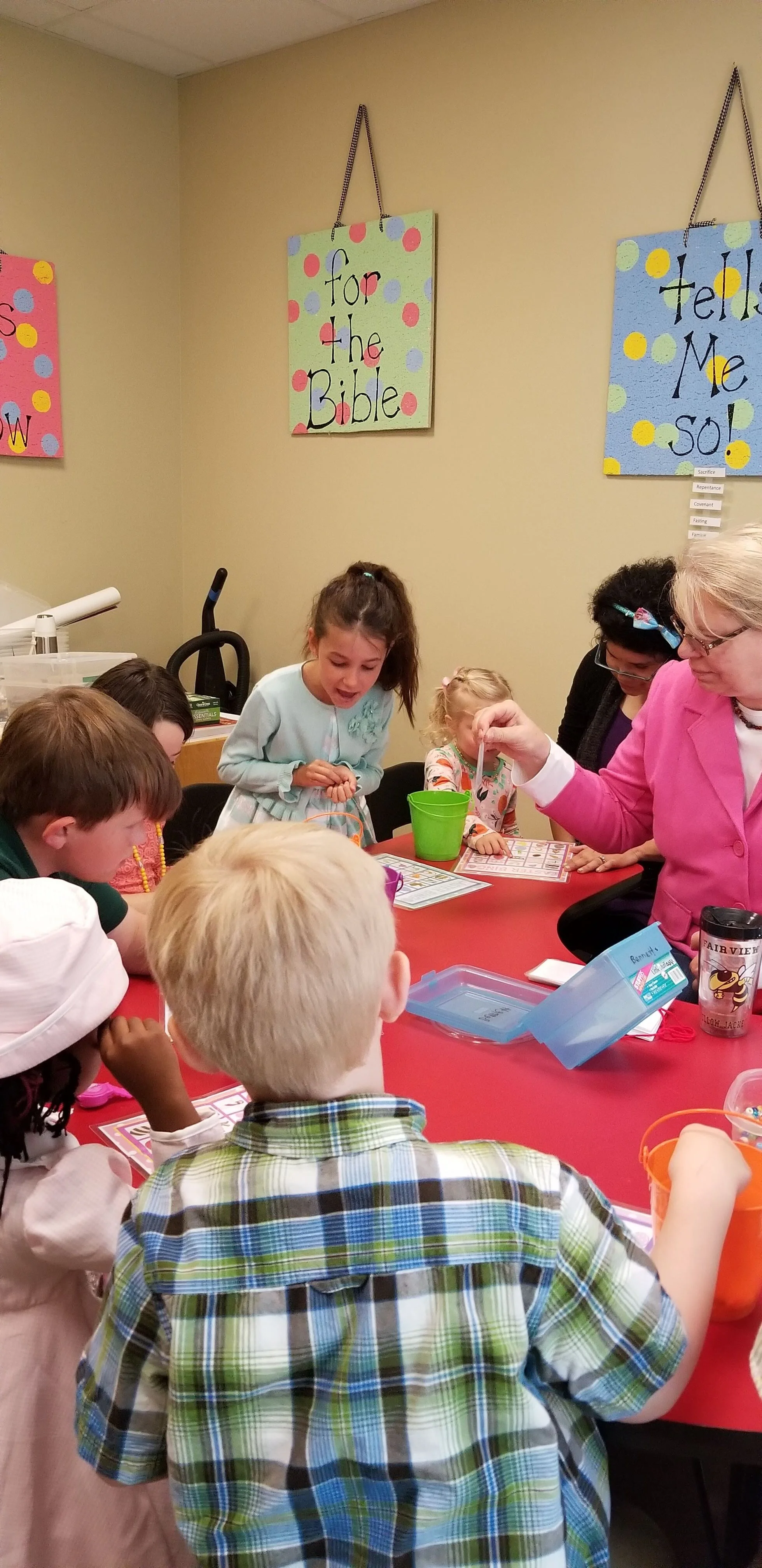 Children and a teacher at a classroom table, engaged in an educational activity. Decorative signs with religious text hang on the wall.