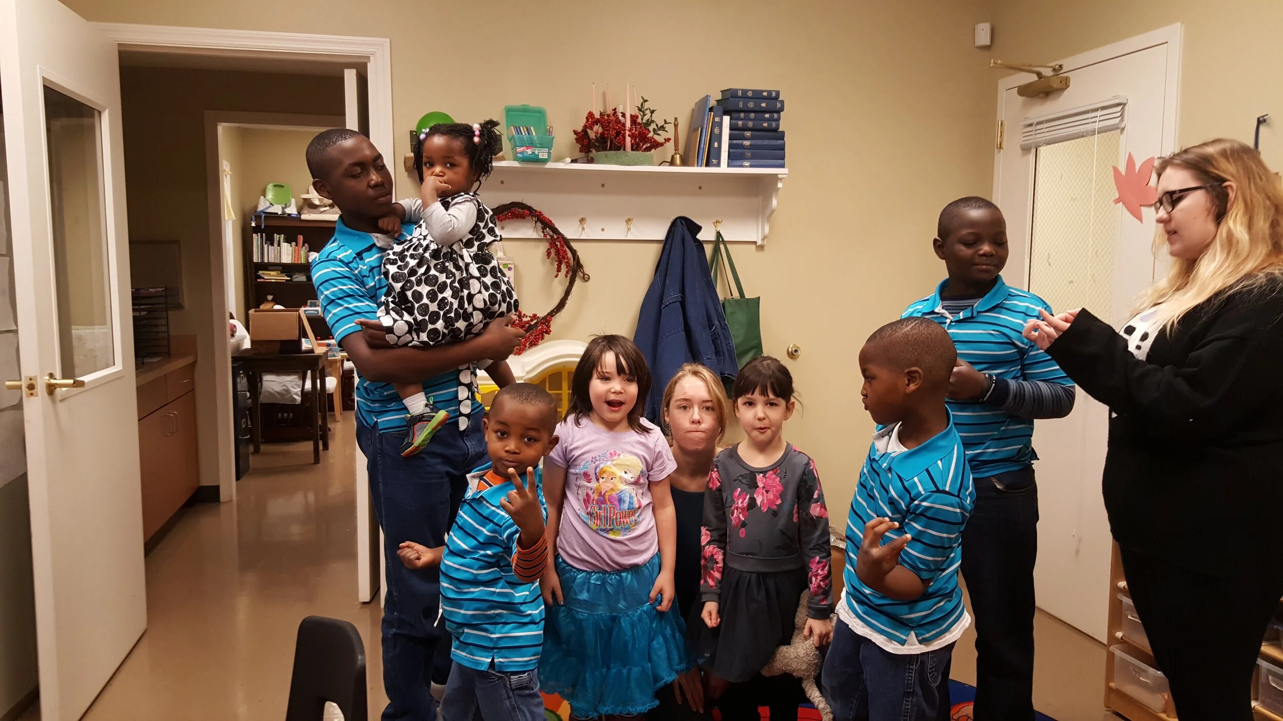 Group of children and two adults in a room with books on a shelf, a door in the background, and coats hanging on hooks.
