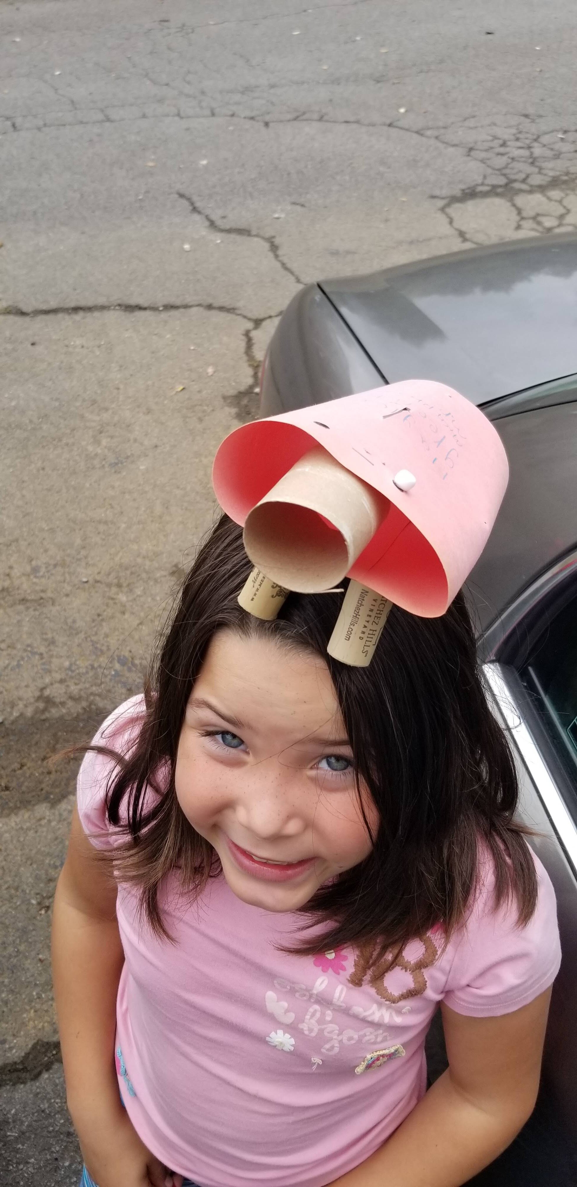 Child wearing a homemade hat made of pink paper and cardboard tubes, standing outside near a car.