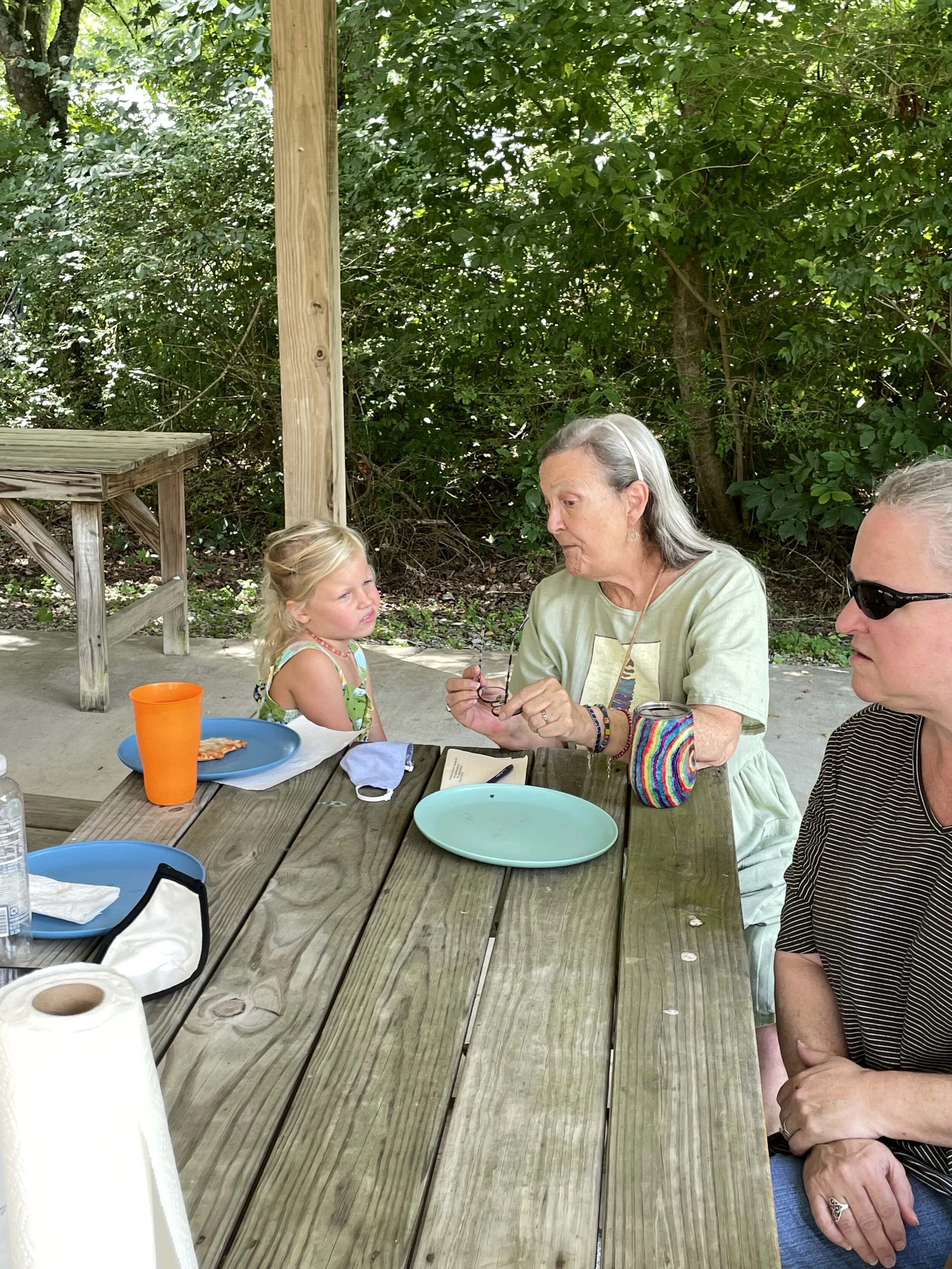 Three people sitting at a wooden picnic table outdoors; a child with a plate and cup, and two adults engaged in conversation.