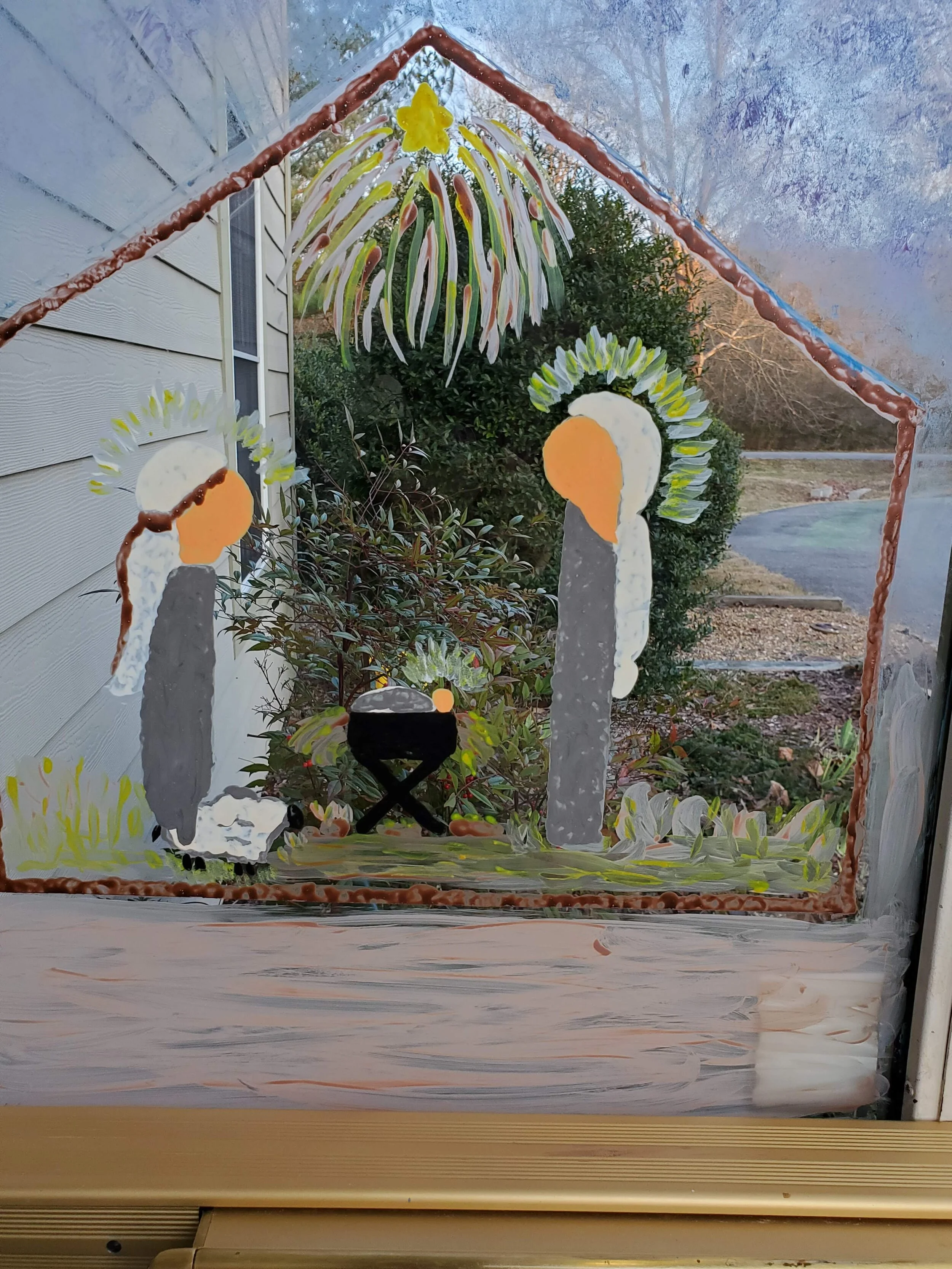 Window painting depicting a simplified Nativity scene with two figures, a manger, and a sheep inside a stable structure, under a starburst pattern.