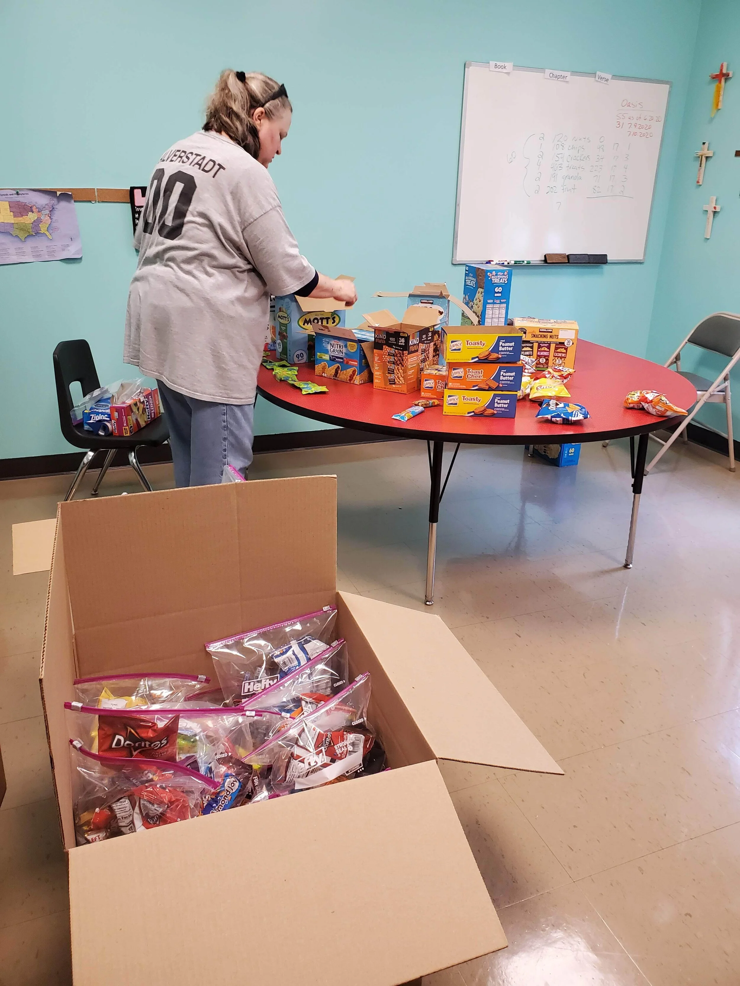 Person organizing snack packages on a table in a classroom, with a large open box containing individual zip-lock snack bags filled with various snacks, including Doritos and granola bars. A whiteboard and chairs are visible in the background.
