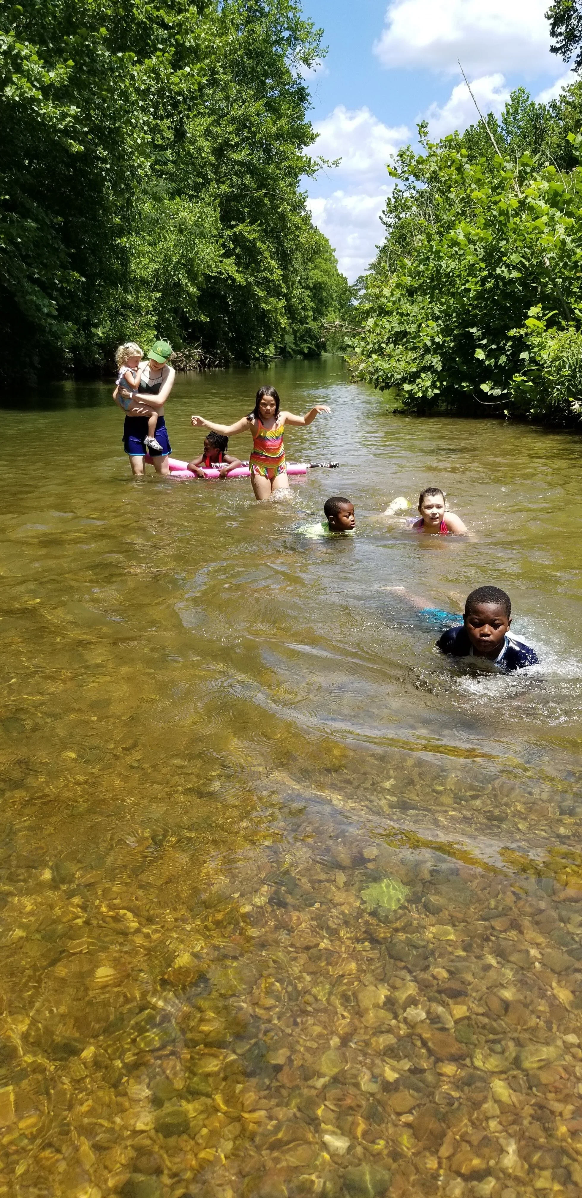 Children and adults playing in a shallow river surrounded by lush green foliage under a blue sky.