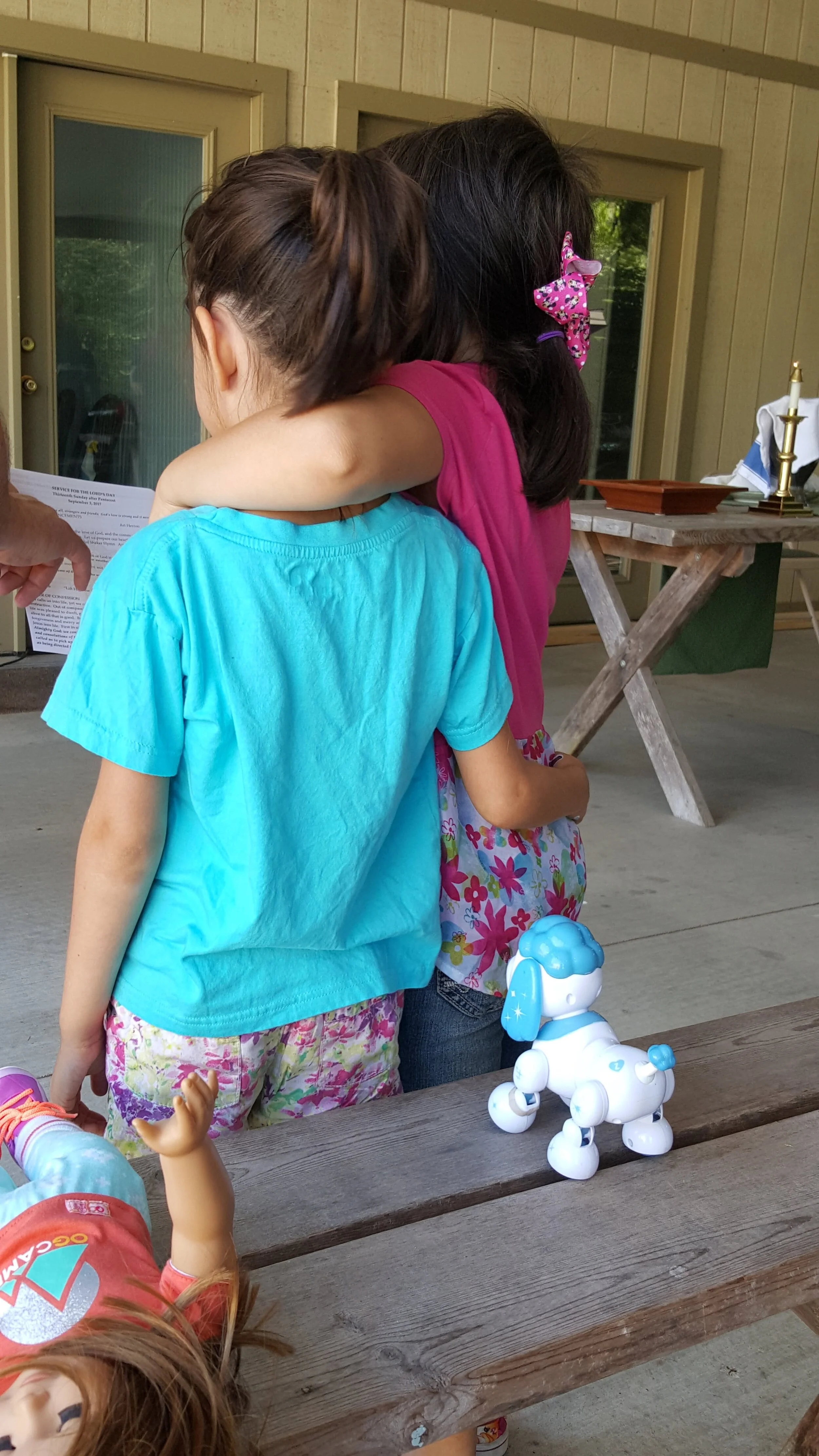 Two children, one wearing a blue shirt and the other a pink one with a large bow hair clip, embrace while facing away. A toy robot dog and a doll are on a wooden table nearby. The setting appears to be an outdoor porch or patio with wooden walls and 