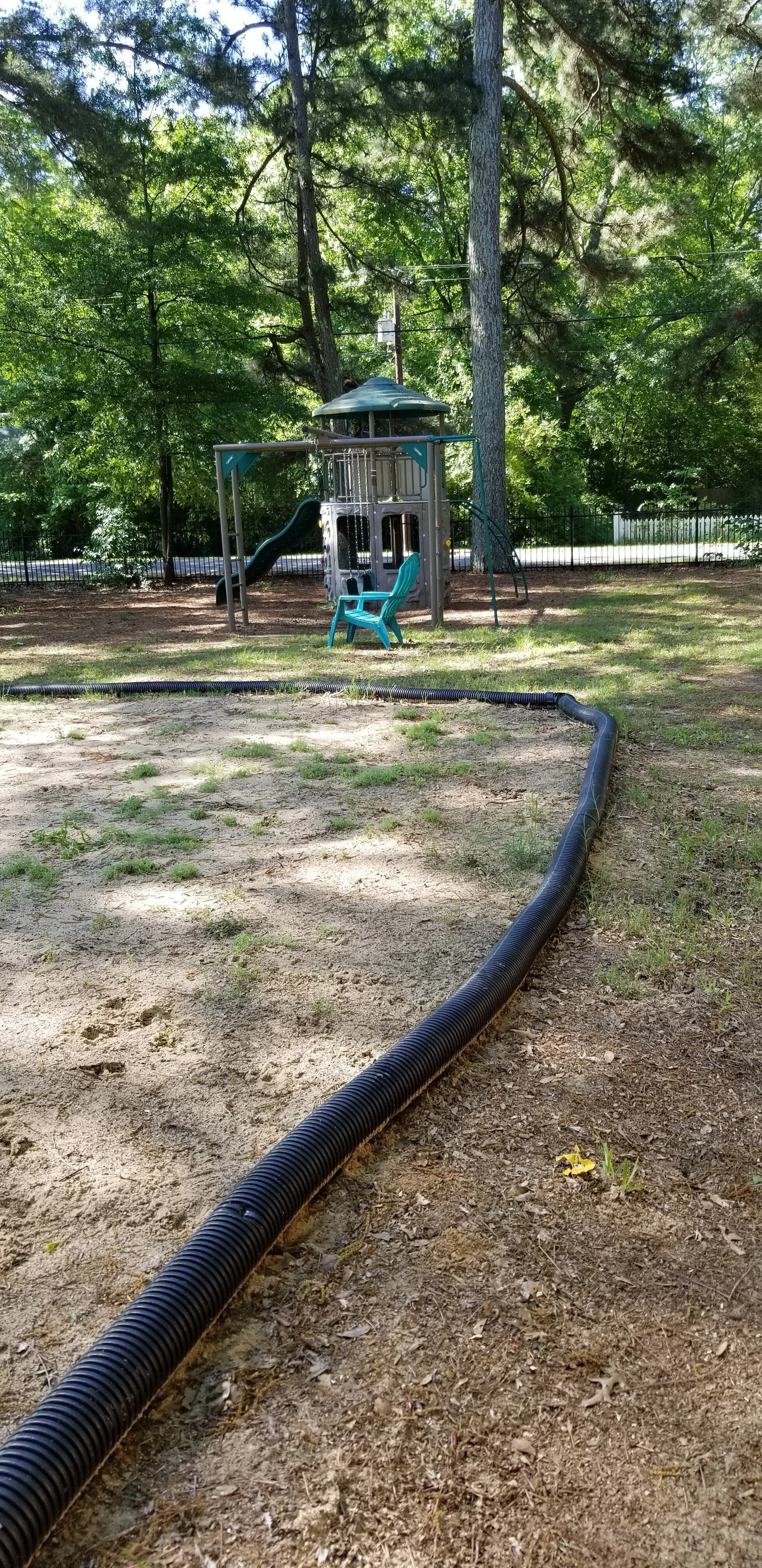Playground with a slide, swings, and a plastic chair surrounded by trees and grass. A black corrugated tube lies on the ground.