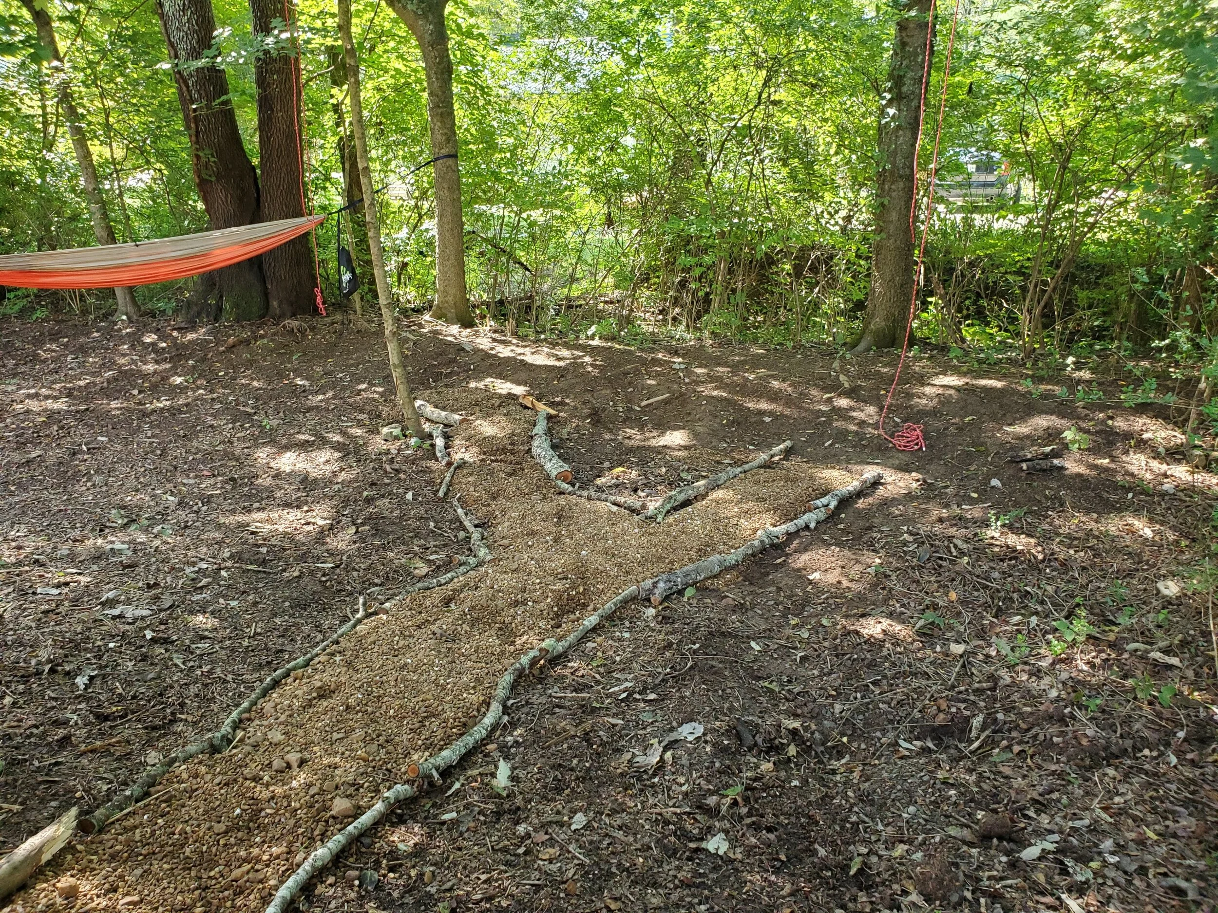 Wooded area with a suspended hammock between trees and a path bordered by sticks and covered with gravel.