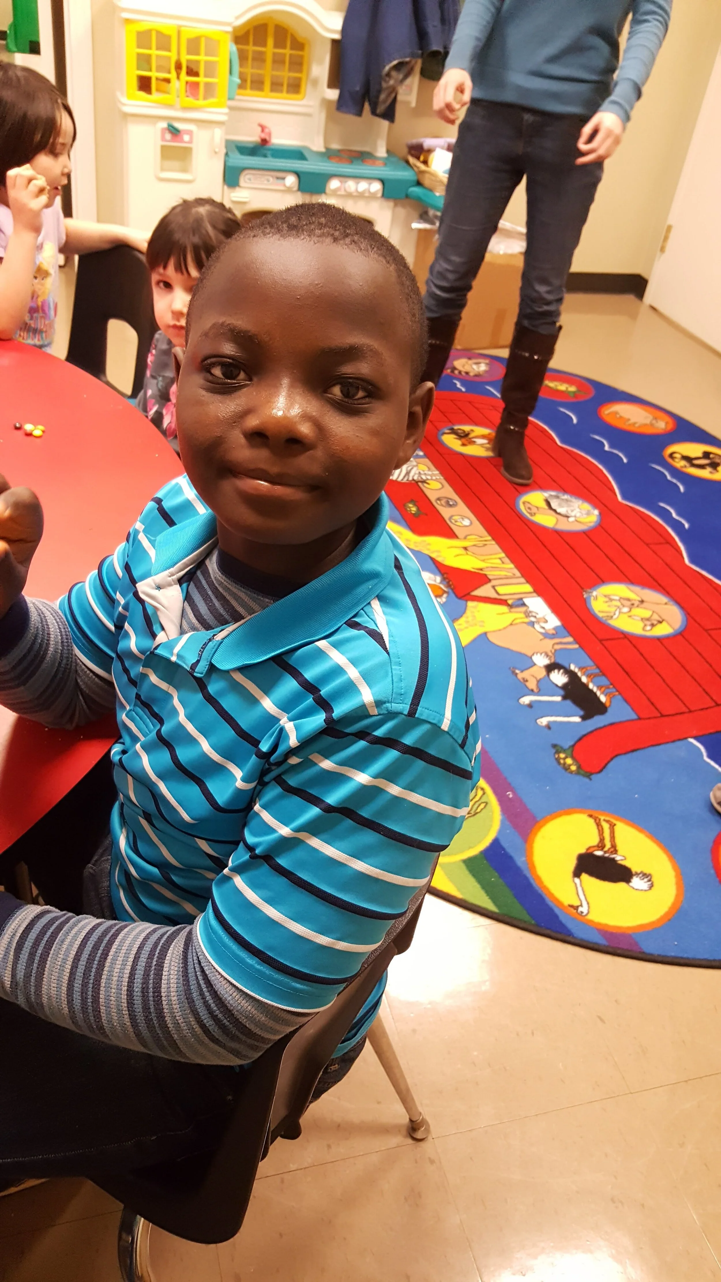 Child in striped shirt at a classroom table, colorful floor mat, play kitchen, and other children in background.