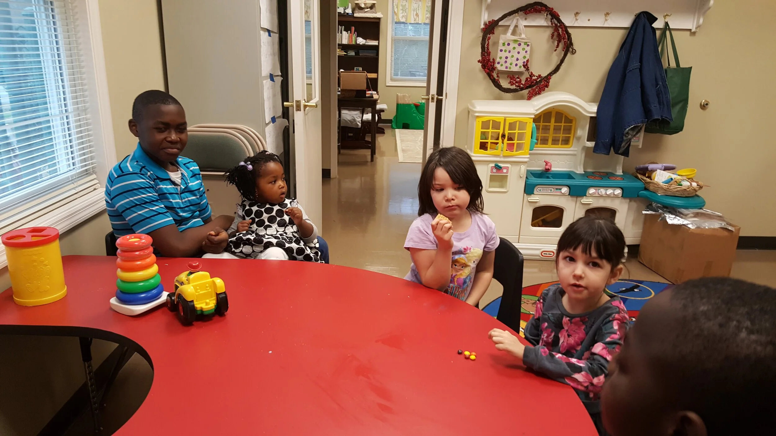 Children sitting around a red table in a playroom with toys, including stacking rings and a toy car. Several children are eating or holding food. A toy kitchen set and coats are visible in the background.
