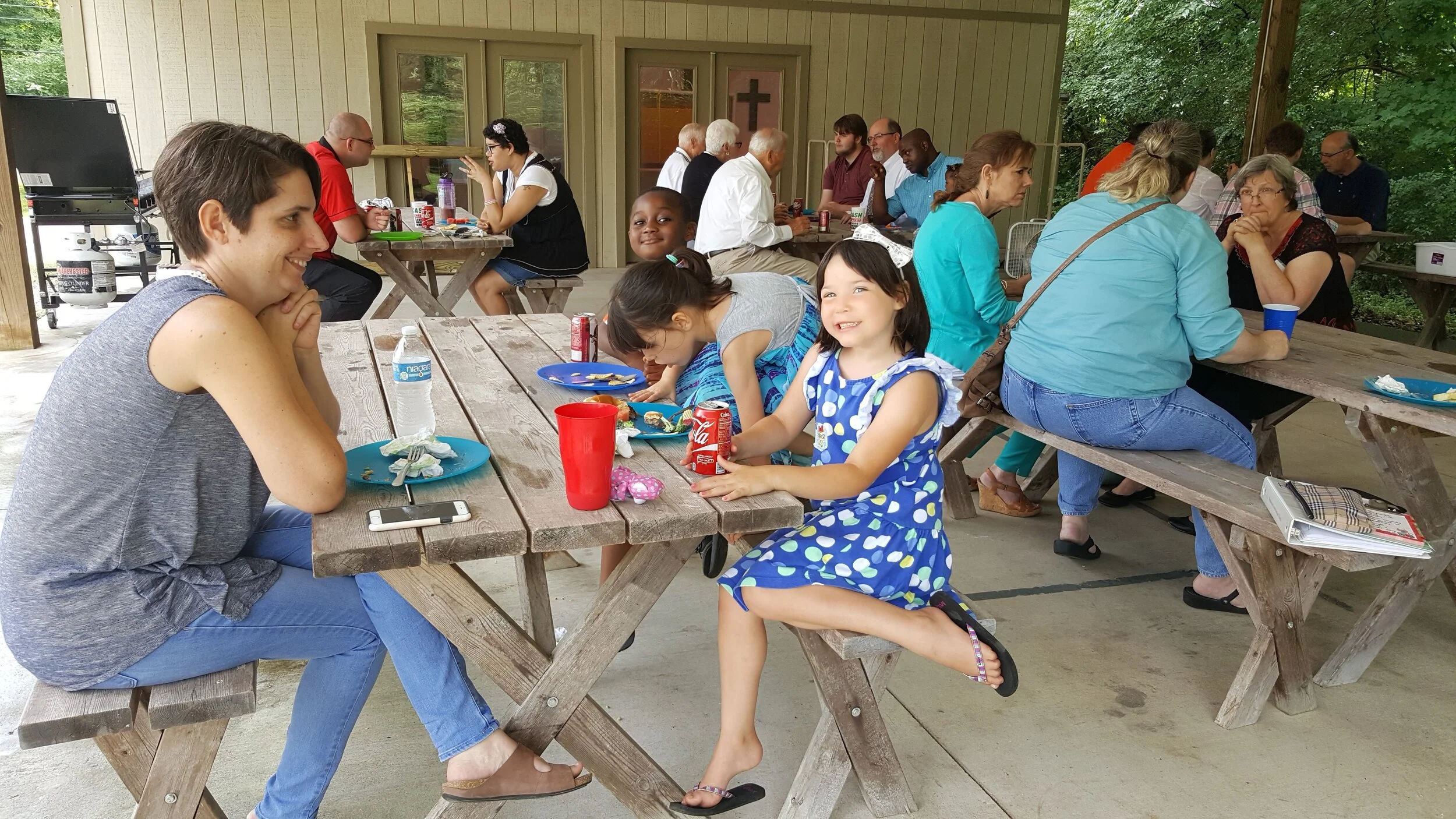 A group of people gathered at a picnic area, sitting at wooden tables. Some are adults, while others are children. They are enjoying food and drinks, with soda cans and plates visible on the tables. The setting is outdoors with a forested background.