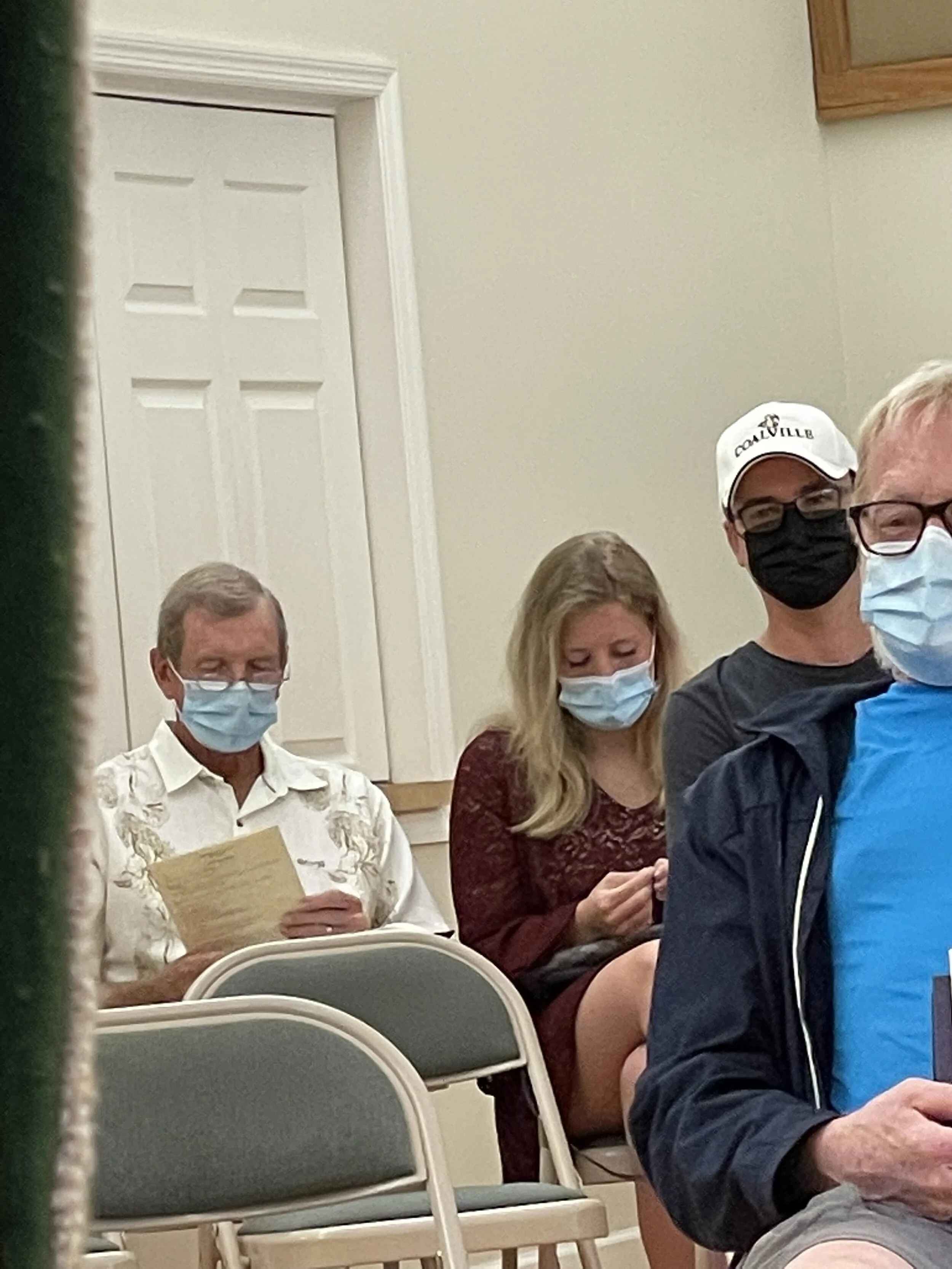 People wearing masks seated indoors, two holding papers, another wearing a white cap, with a closed door in the background.