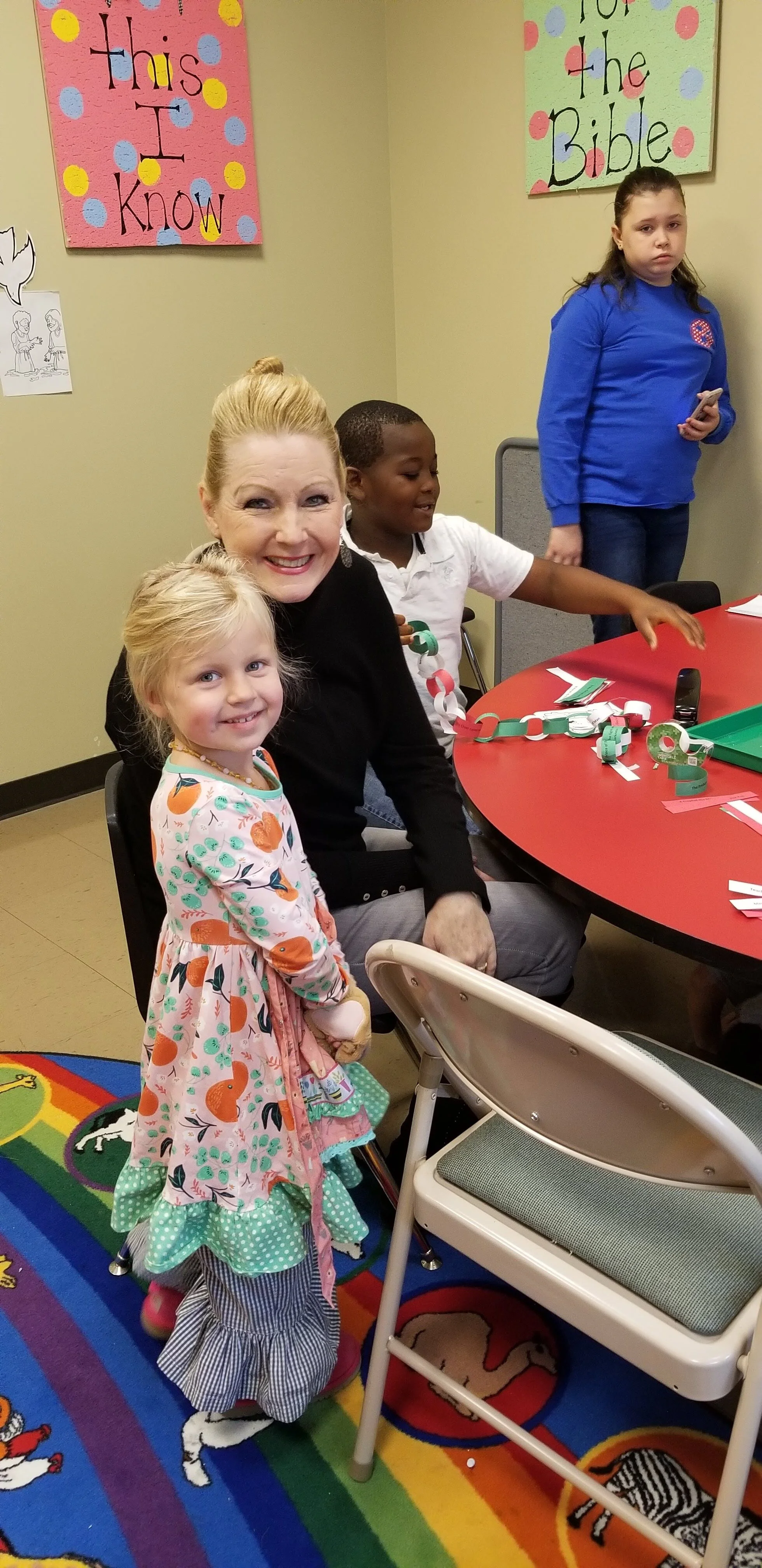 Group of people, including a smiling child and adult, at a table with crafts and paper chains, in a colorful room with educational posters on the walls.