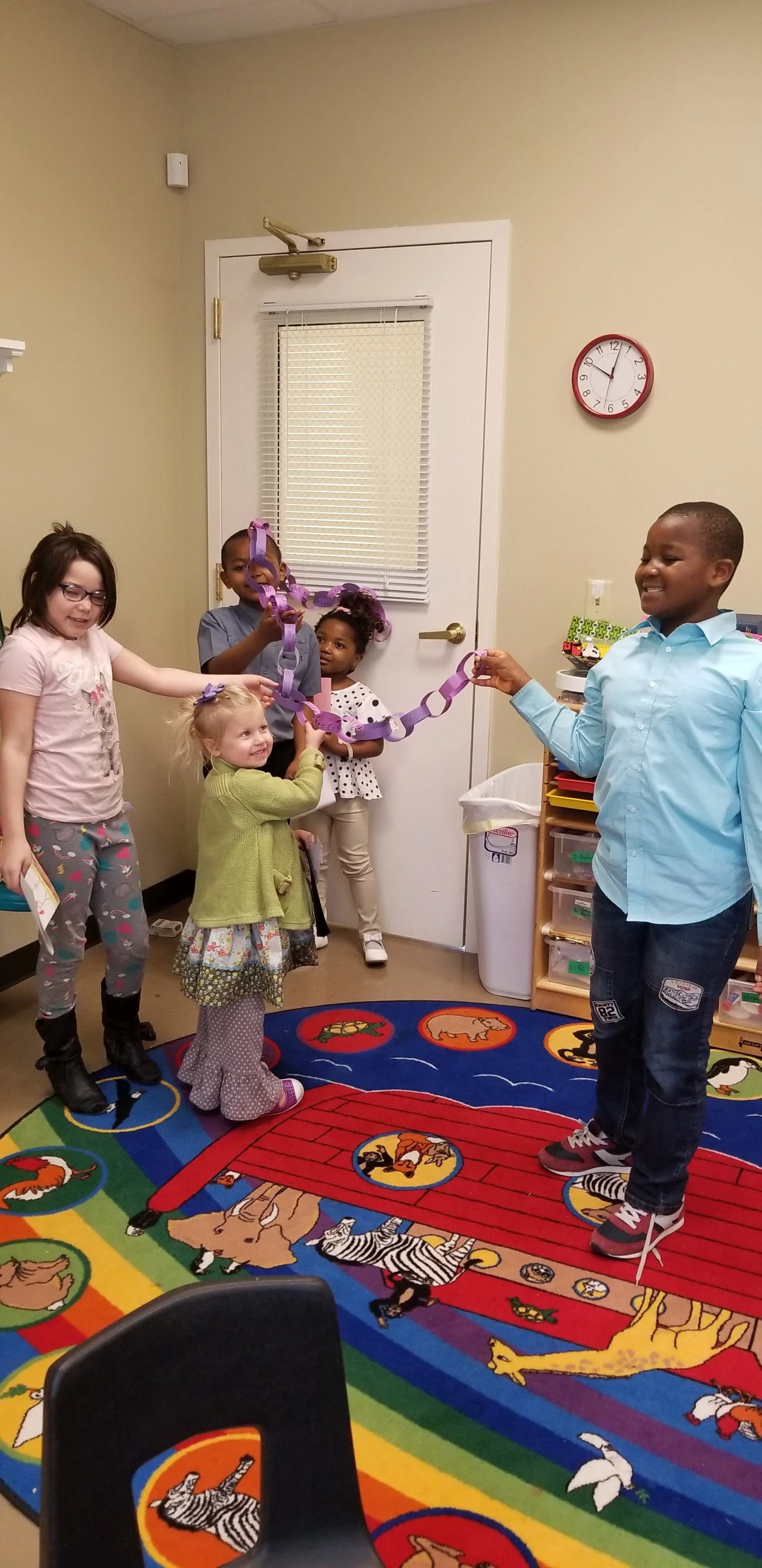 Children in a classroom holding a purple paper chain, standing on a colorful animal-themed rug.
