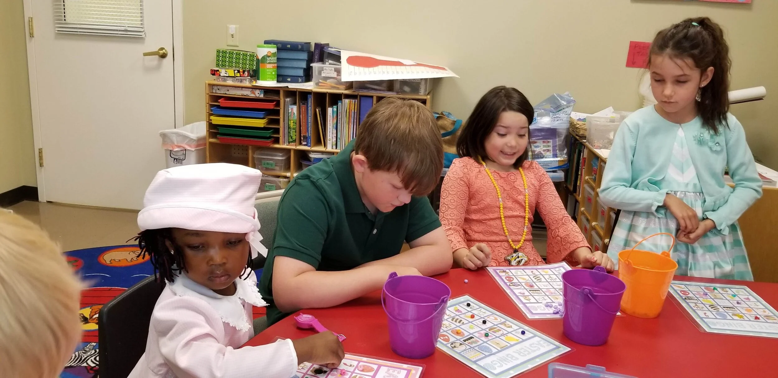 Children playing an educational game at a table in a classroom setting, with colorful buckets and game boards on the table.