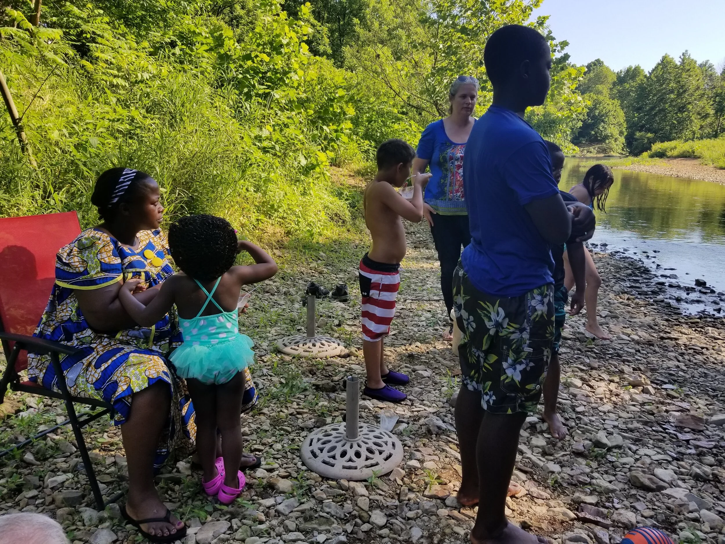People standing and sitting by a riverbank, surrounded by greenery, on a sunny day.
