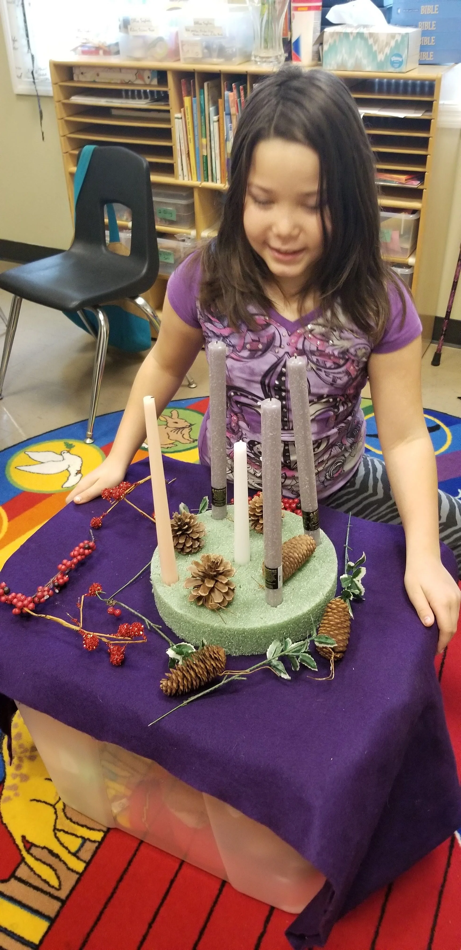 Child smiling while arranging holiday-themed candles with pine cones and red berries on a purple tablecloth, in a classroom setting.