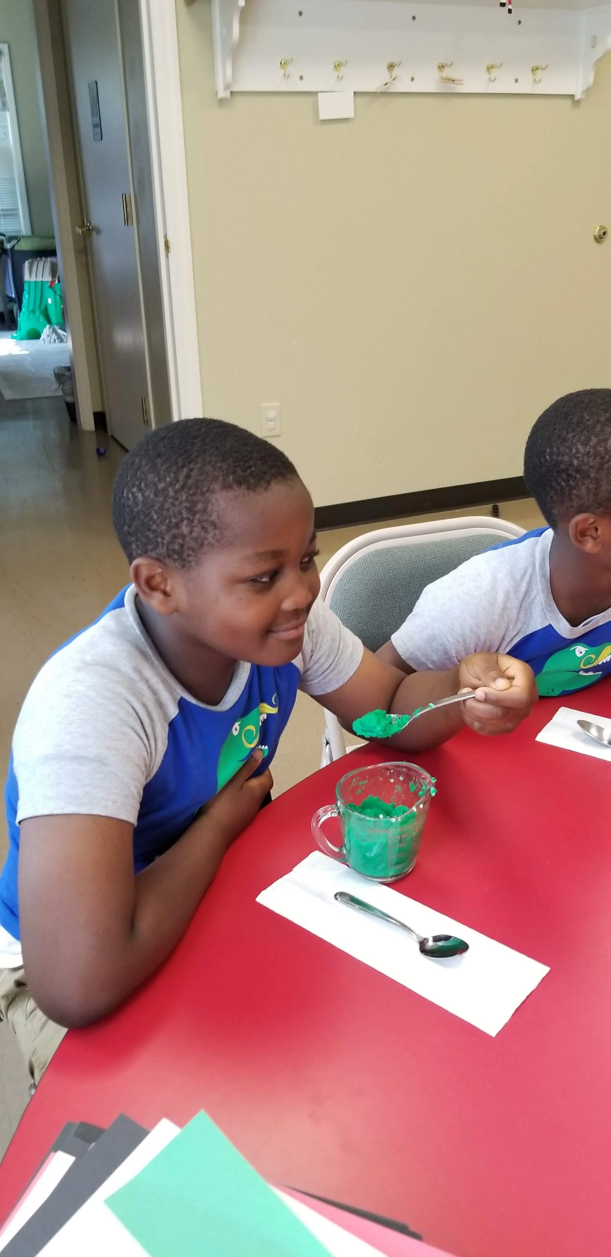 Two children sitting at a red table, one holding a spoon with green liquid from a measuring cup.