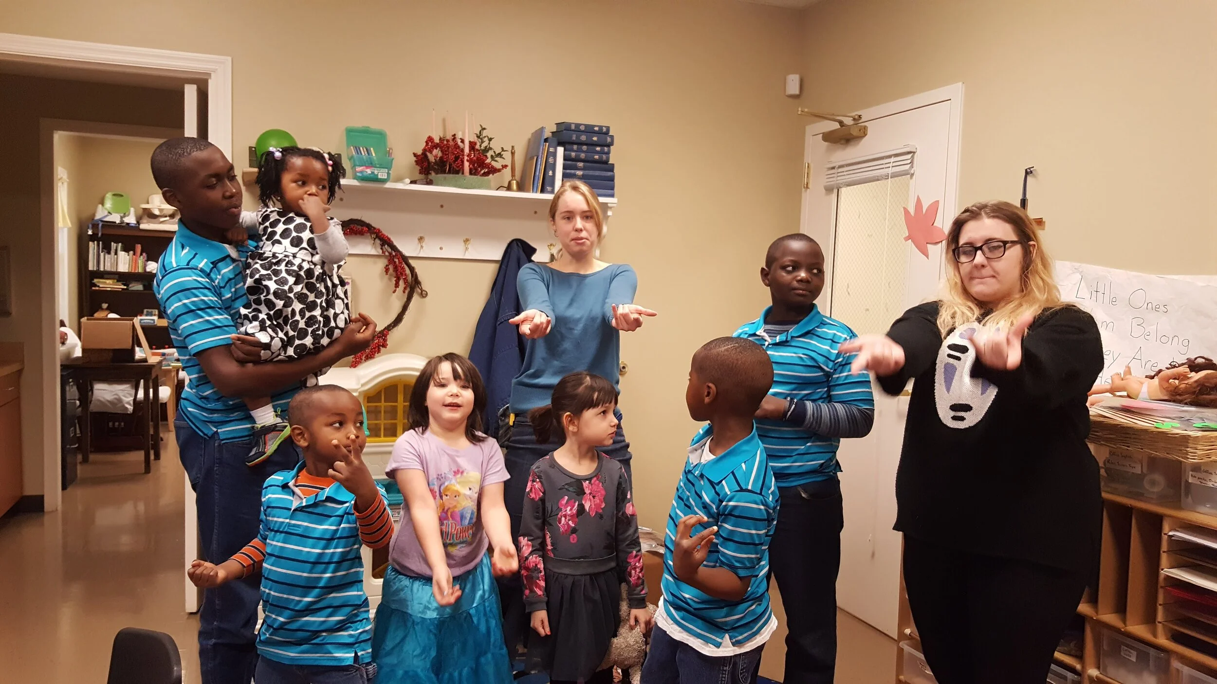 A group of children and adults in a classroom, some gesturing with their hands.