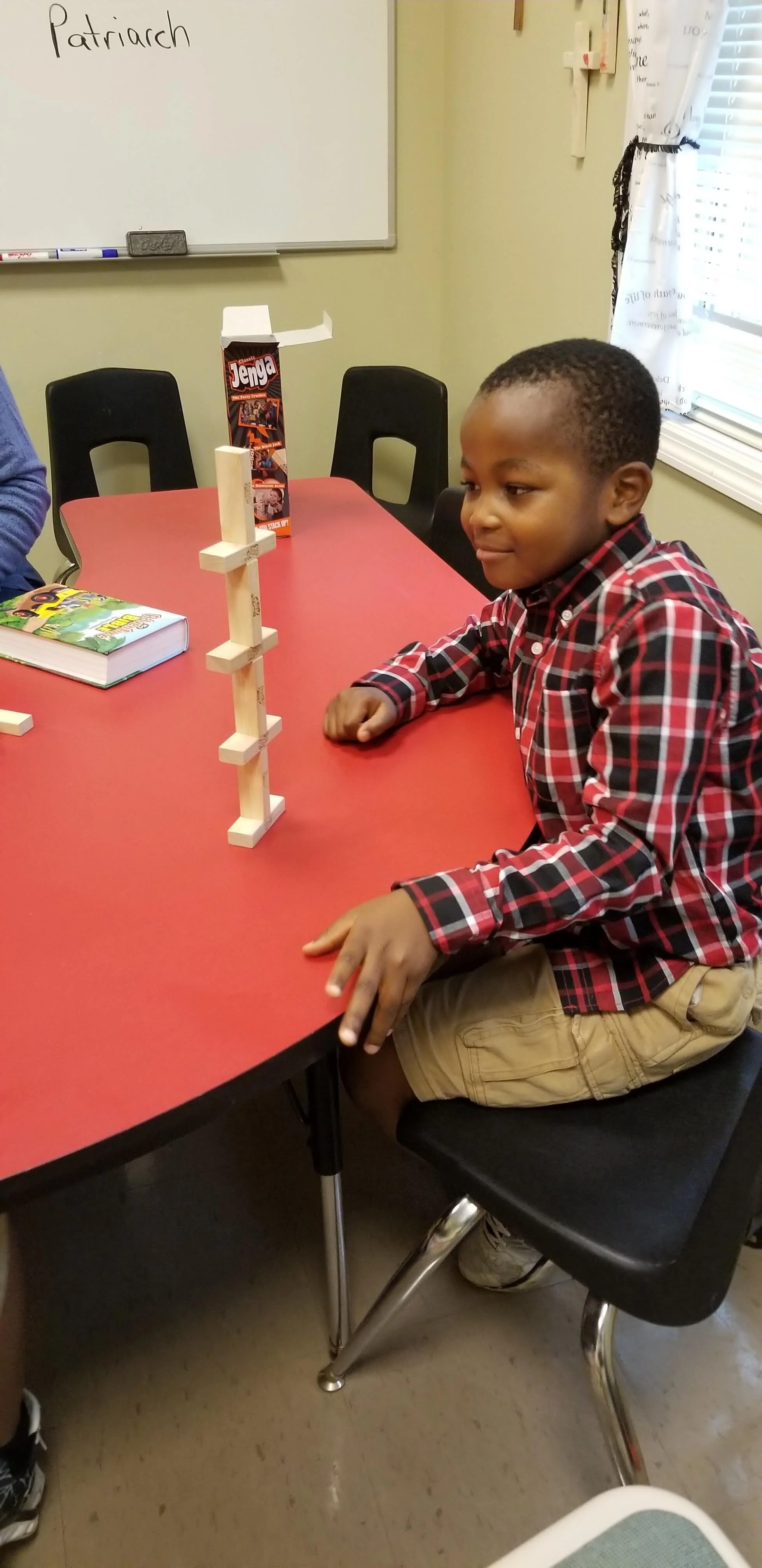 Child playing with Jenga blocks on a red table, with a Jenga box and book visible.