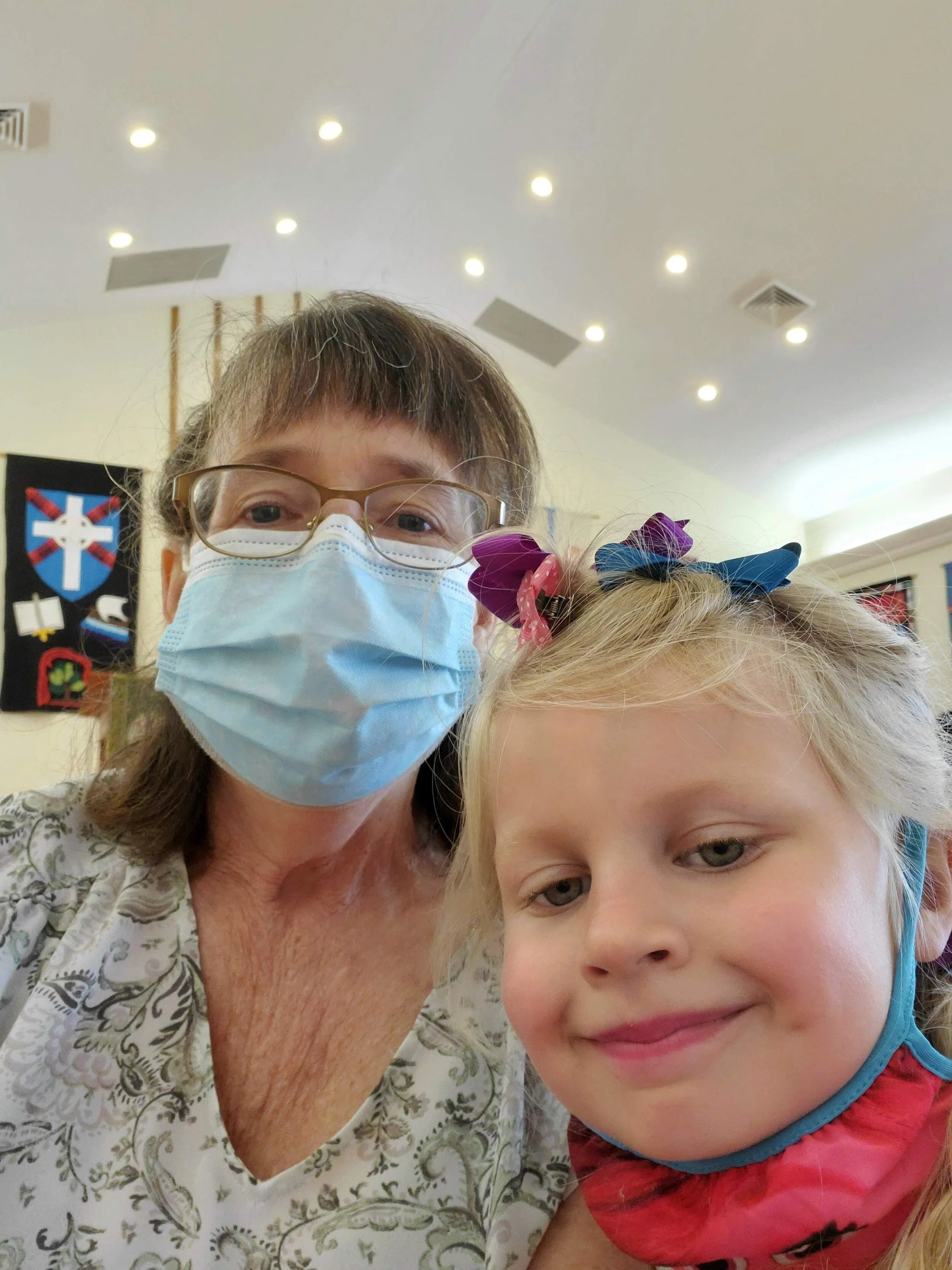 A woman wearing glasses and a blue face mask sits next to a child with blonde hair, wearing colorful hair ties and a lowered face mask. They appear to be in an indoor setting with bright lights.