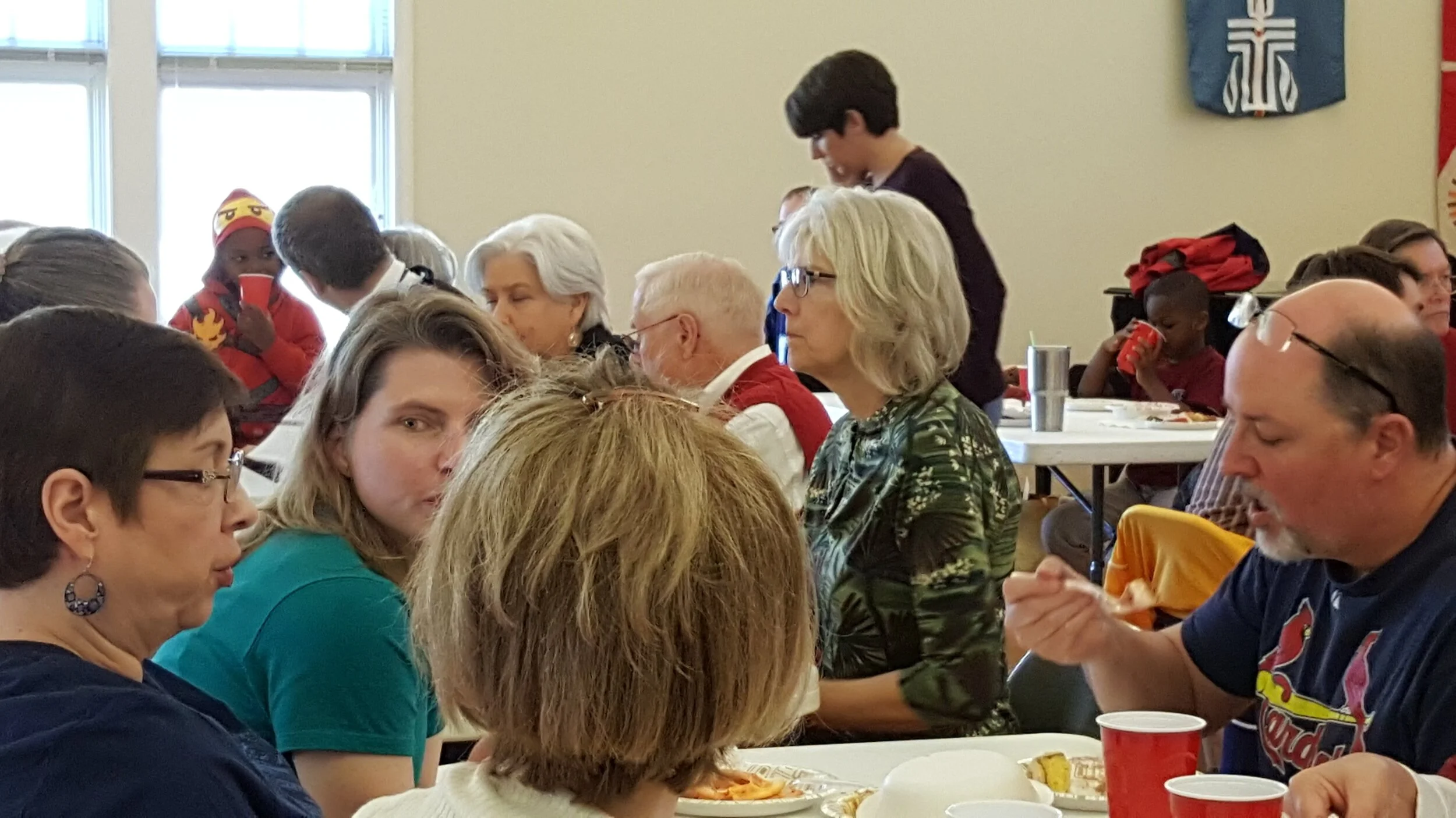 People sitting at tables, eating and drinking at a communal gathering in a brightly lit room.