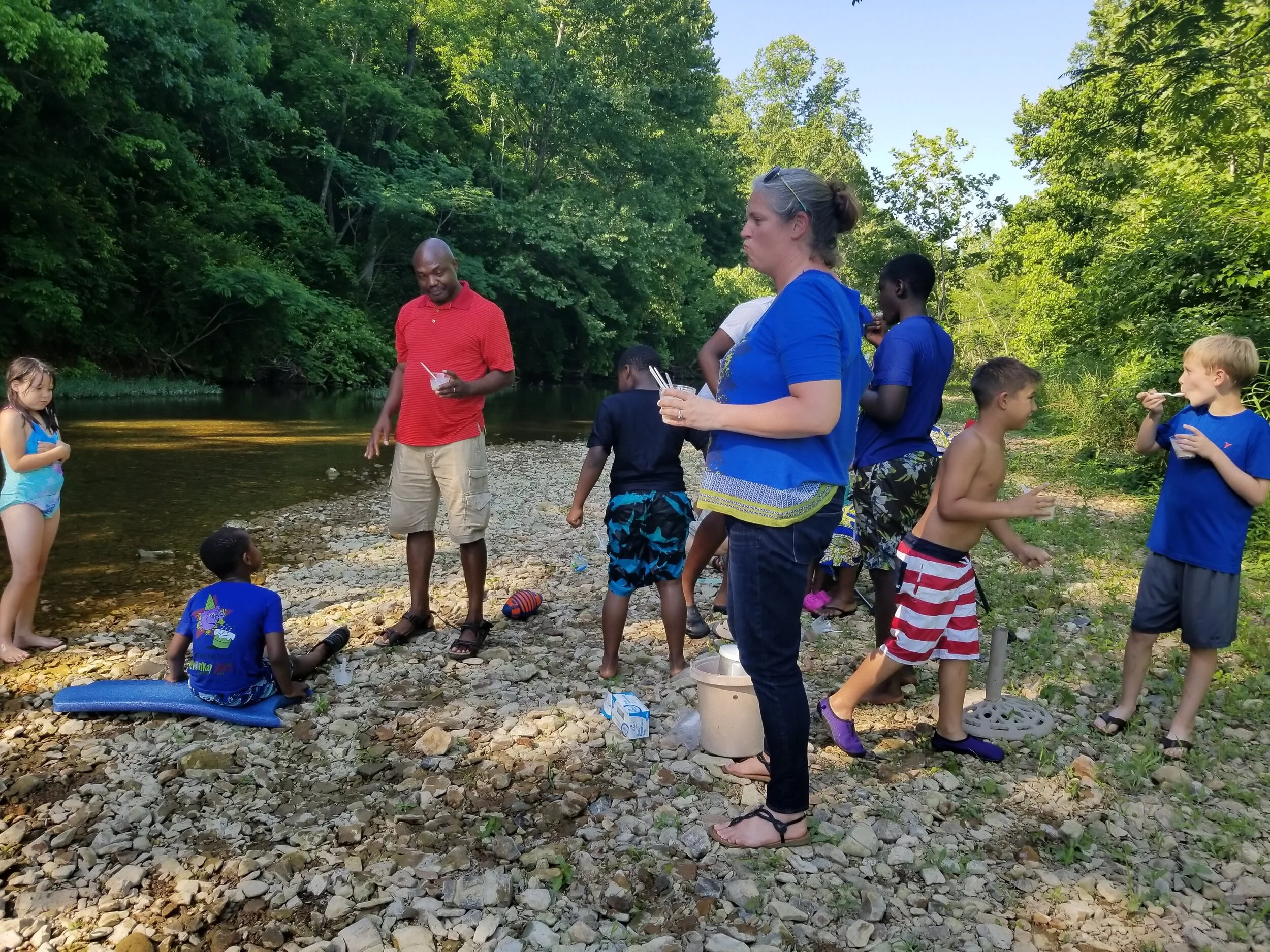 Group of people, including adults and children, enjoying a sunny day by a riverside with rocks, surrounded by greenery.