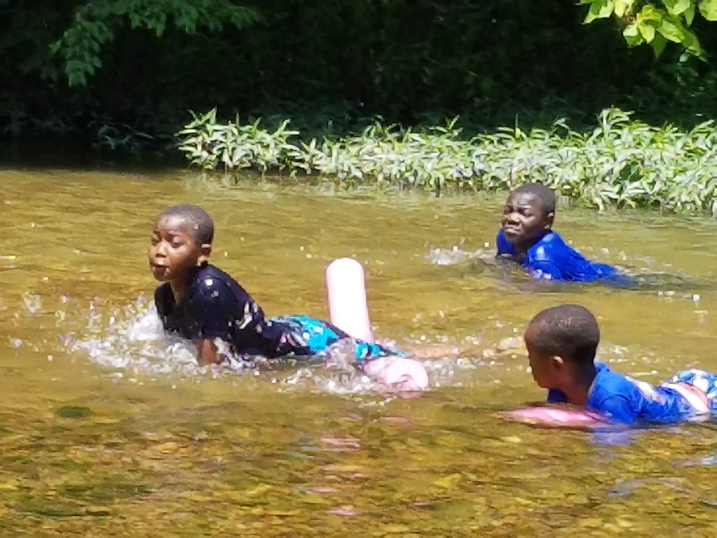 Three children playing in a shallow creek with inflatable toys, surrounded by greenery.