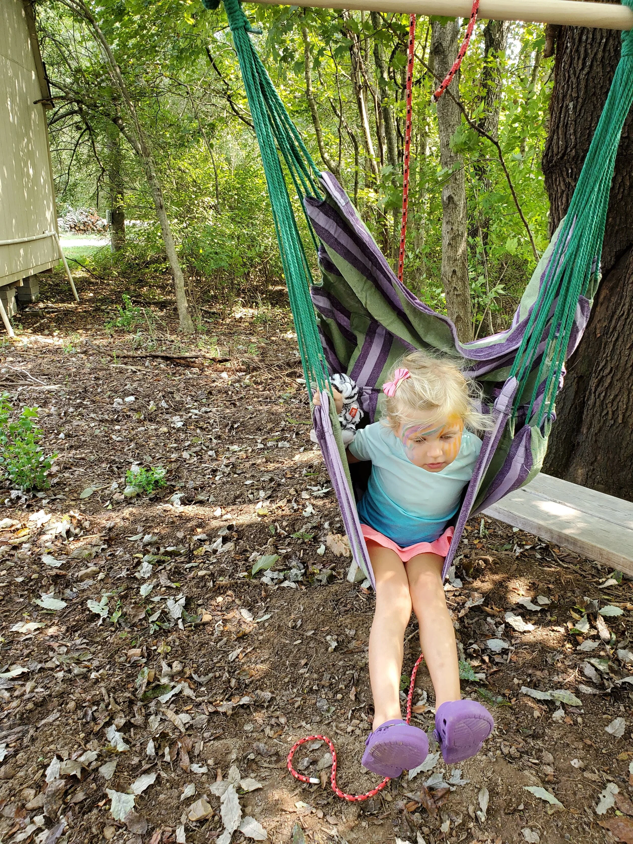 A child with face paint sits in a striped hammock chair outdoors, surrounded by trees and leaf-covered ground.