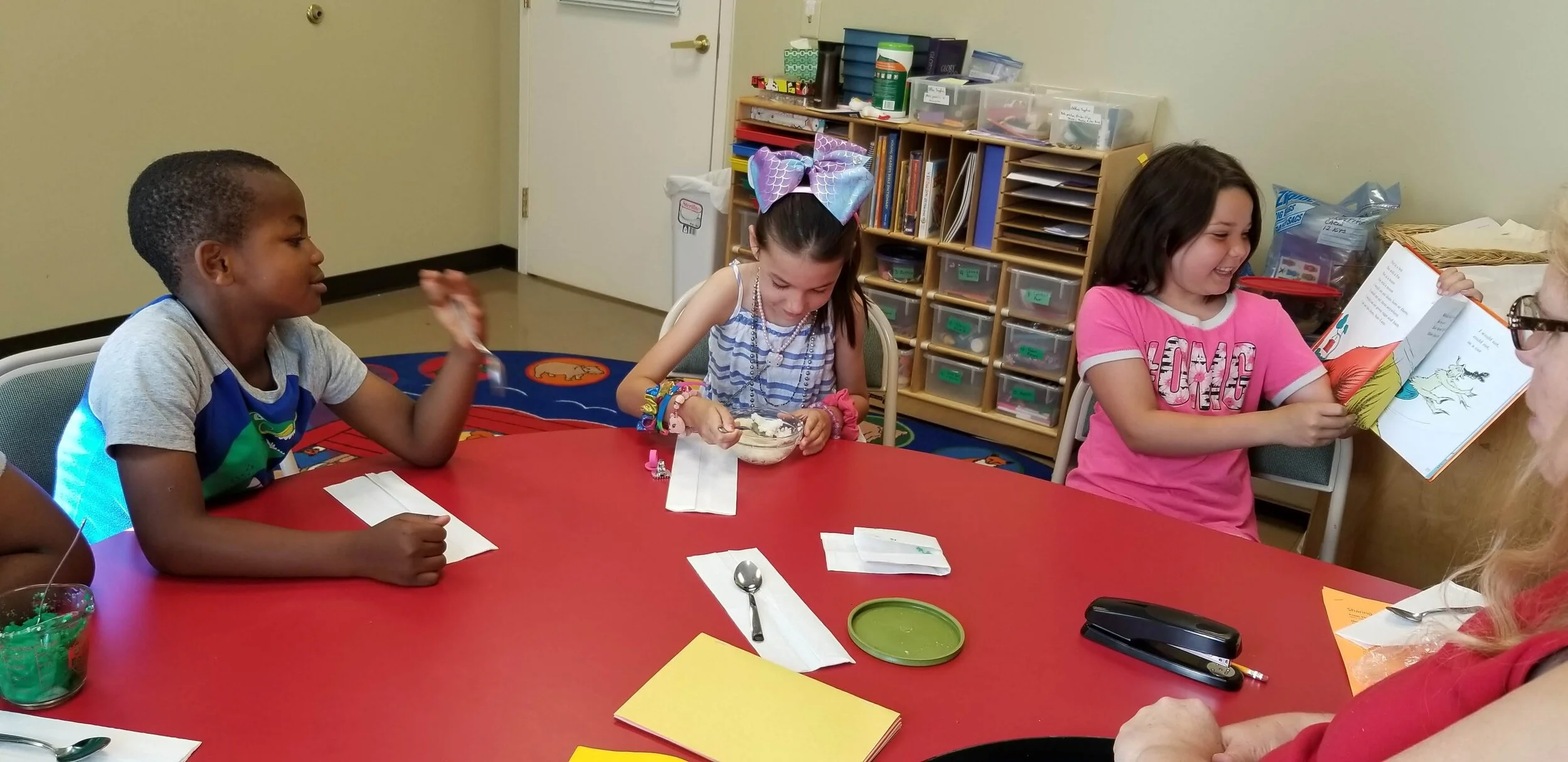 Three children sitting around a red table in a classroom. One child is reading a book, another is eating from a bowl, and the third is looking on. The background has shelves with books and storage boxes.