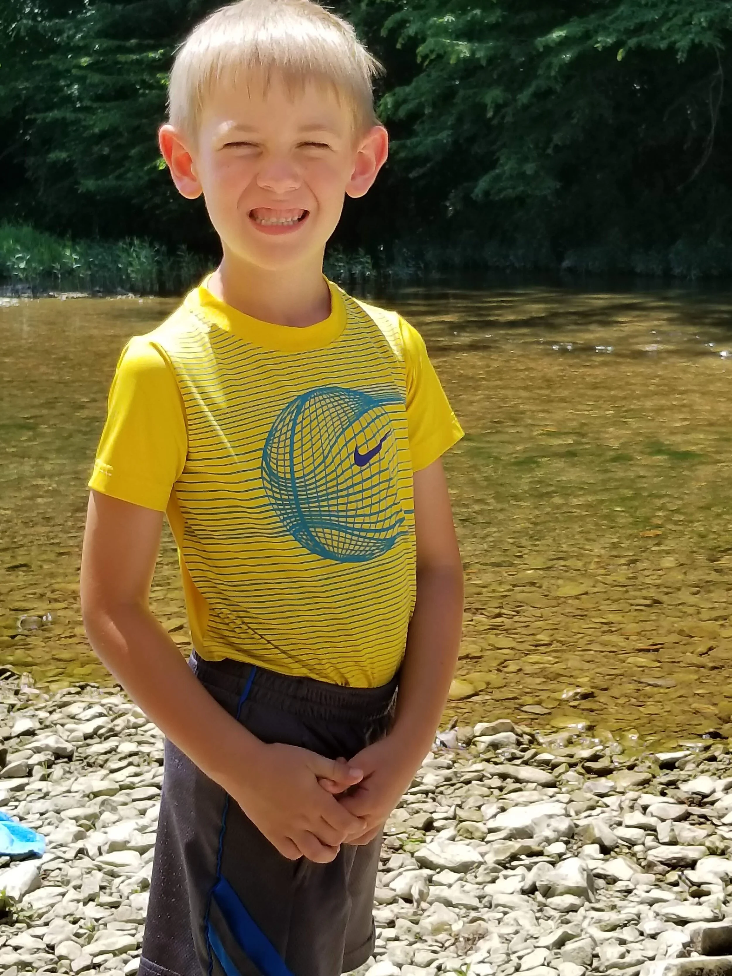 Smiling child in a yellow shirt standing by a rocky stream with trees in the background.
