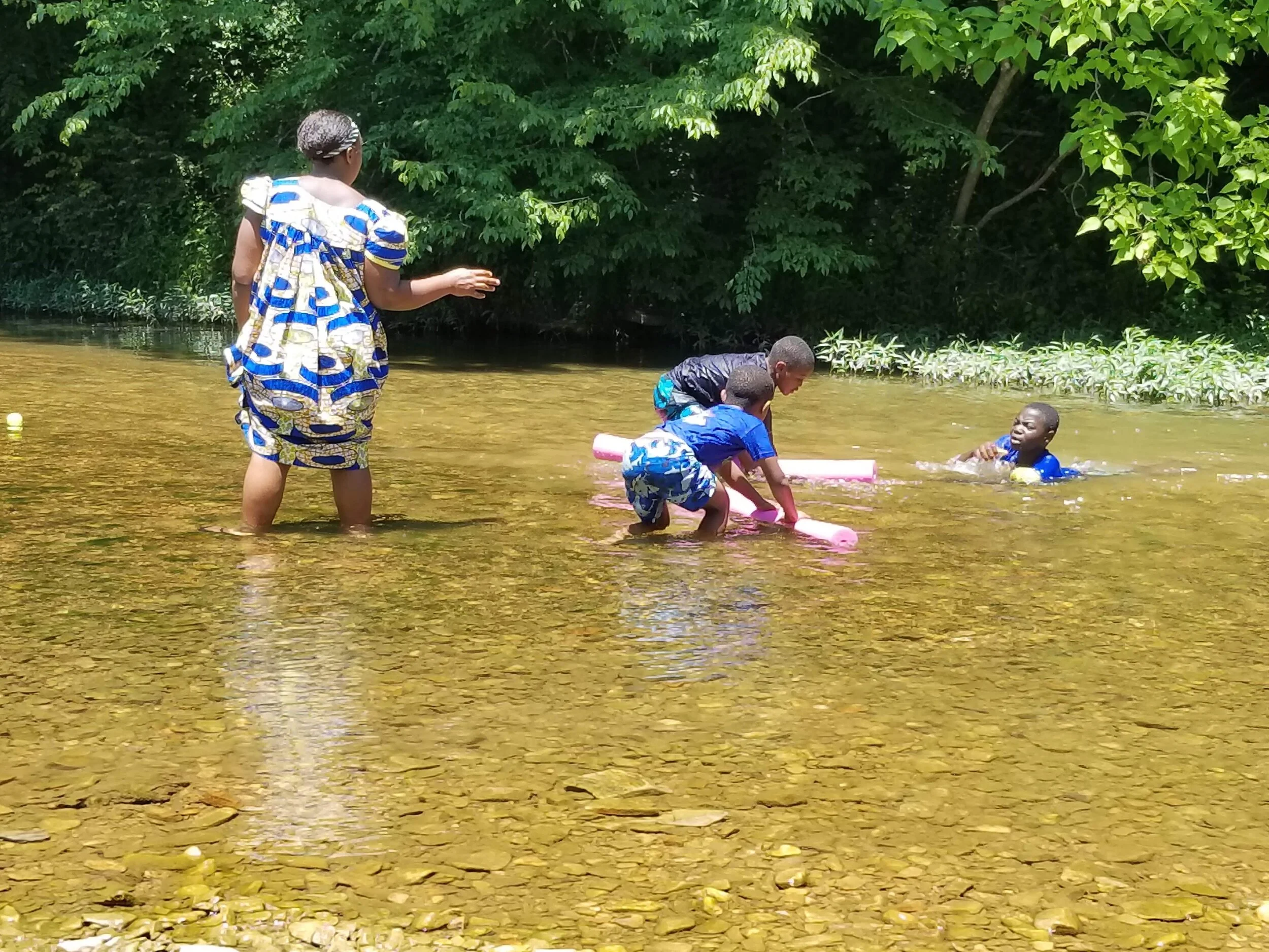 Children and adult playing in a shallow river with trees in background.