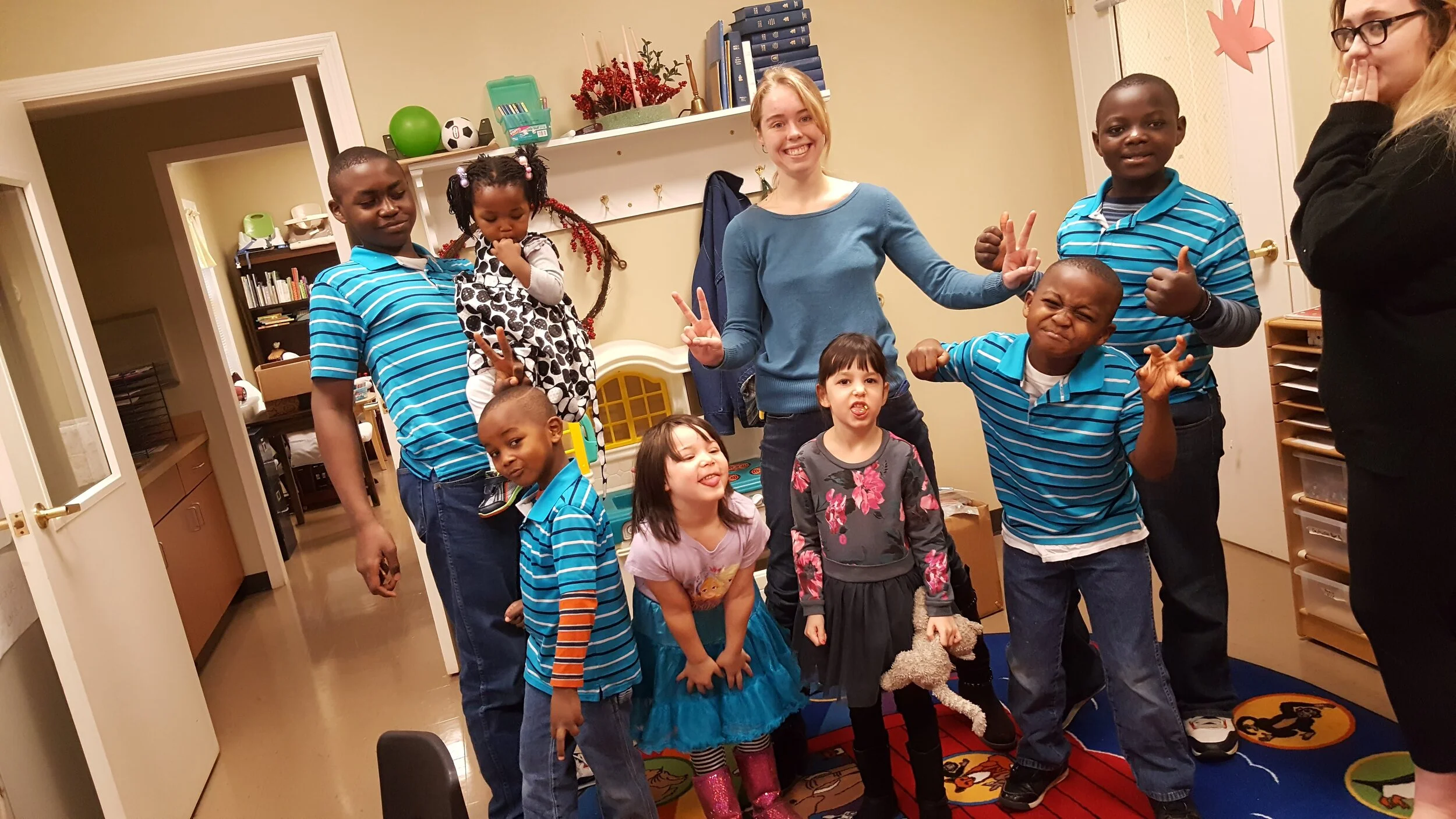 Group of children and two adults posing playfully in a room with books and children's toys.