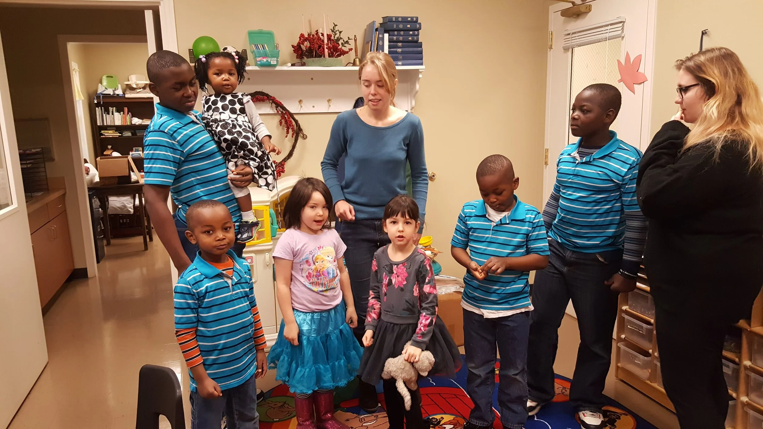 A group of children and two adults standing in a room with educational materials on shelves. Some children are wearing striped shirts and two girls are wearing colorful dresses. An adult is holding a small child. The room has a cozy and educational a