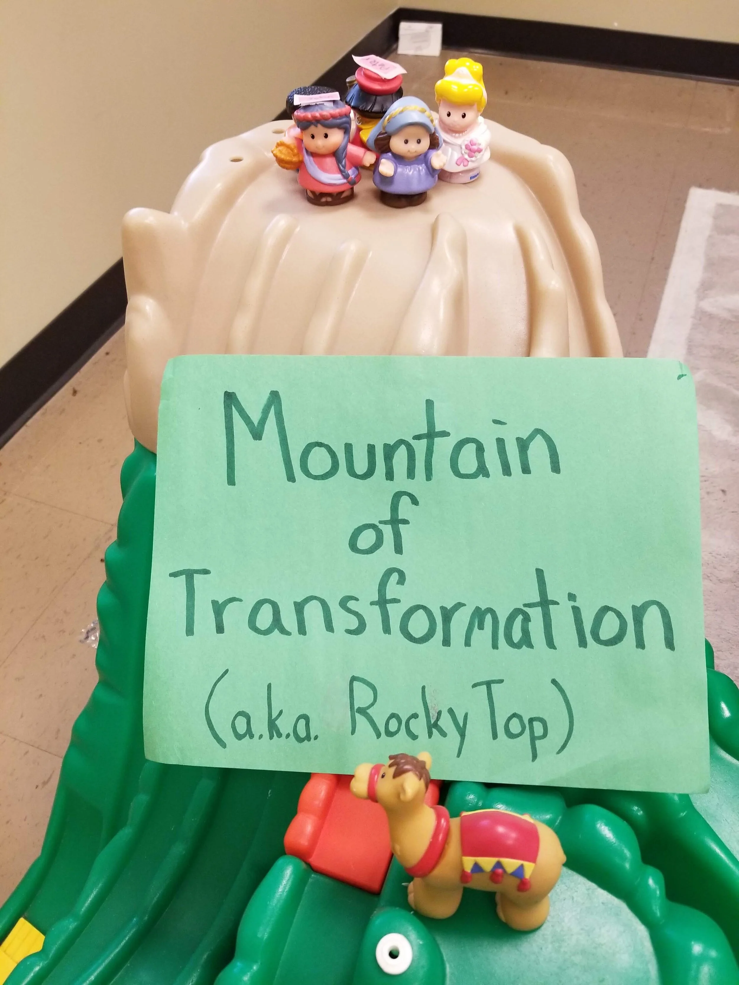Plastic toy figures on a play mountain with a sign reading "Mountain of Transformation (a.k.a. Rocky Top)" and a toy camel nearby.
