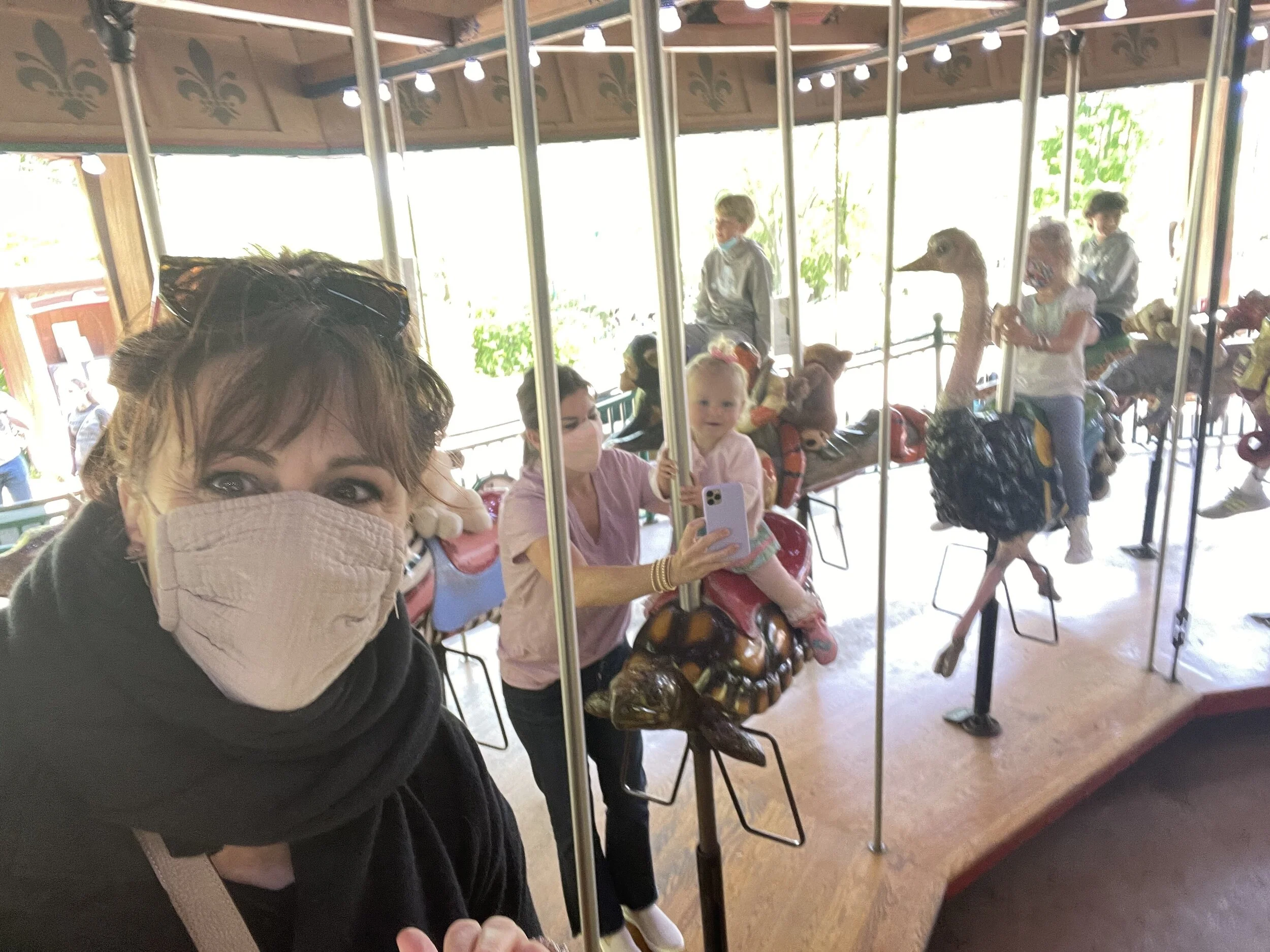 People riding a carousel, including adults and children, with face masks. A child is sitting on an animal ride, while an adult holds a smartphone for a selfie. The carousel roof has decorative elements and lights.